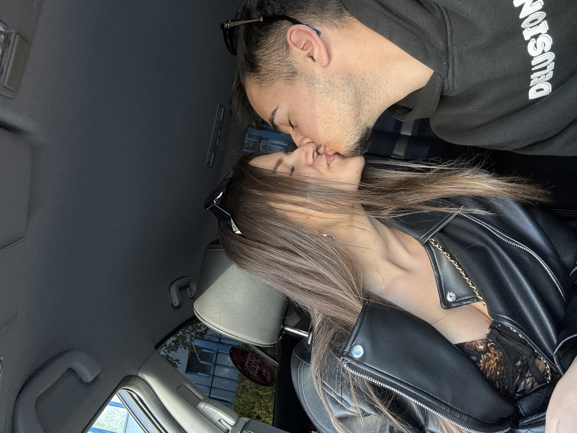 A man and a woman are kissing inside a vehicle. The man has short dark hair, a beard, and is wearing sunglasses on his head and a black sweatshirt with white lettering that reads "Delusion". The woman has long brown hair, is wearing sunglasses on her head, a black leather jacket, and a lace top. The interior of the vehicle is gray. A window is visible in the background, showing a building and trees outside. The car appears to be parked. I am unable to provide the exact location in the city of Sofia, Bulgaria, with the provided information.