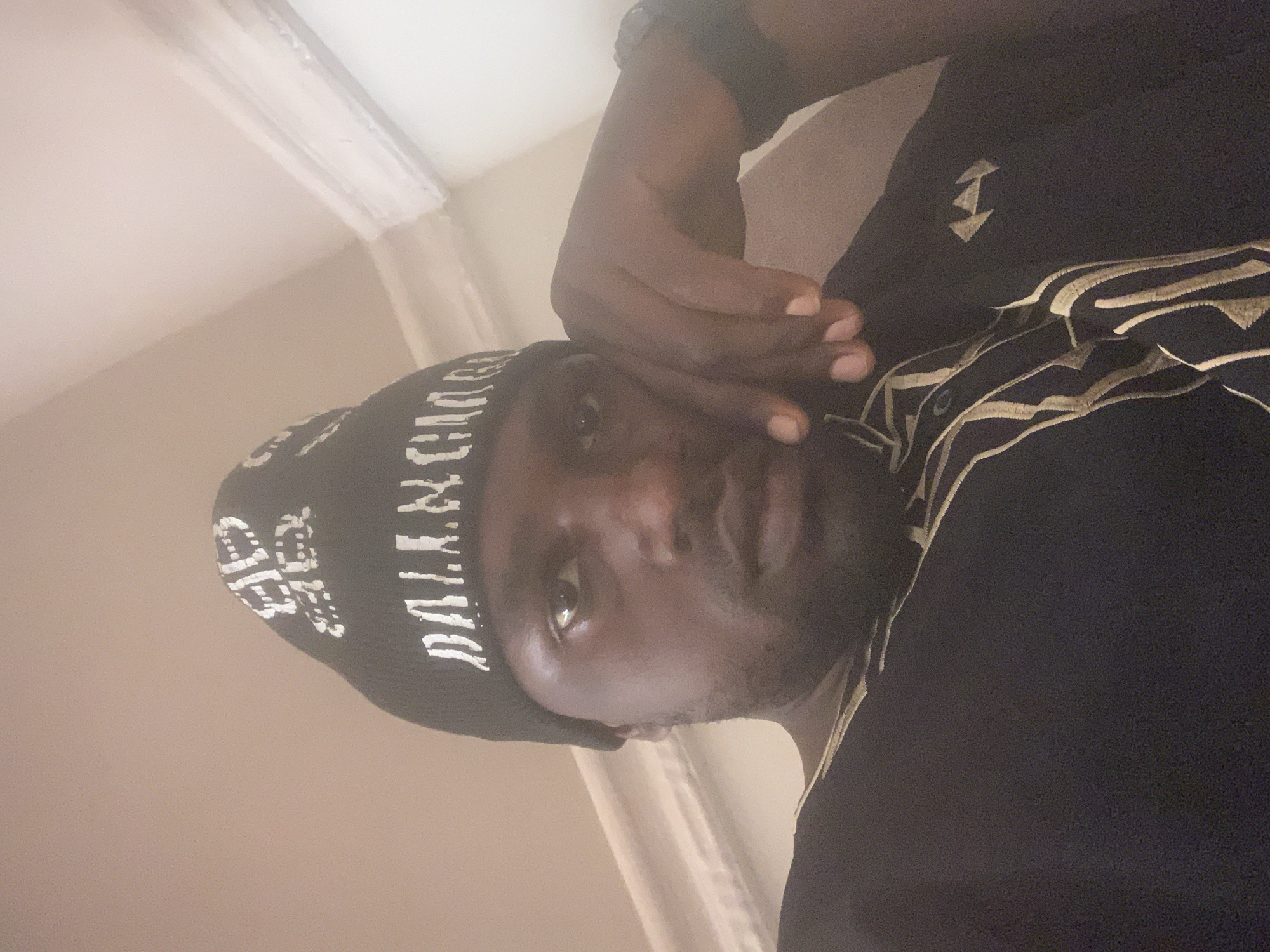 The image is a close-up, selfie-style photograph of a Black man wearing a black beanie with white lettering that appears to spell "BALENCIAGA". He is also wearing a dark garment with gold embroidery. His right hand is resting on his cheek, and his left arm is not visible. The background appears to be an indoor setting with a plain, light-colored wall and some architectural molding. The lighting is soft and appears to be coming from above. There is no indication of the location being in Gura Top, Nigeria.