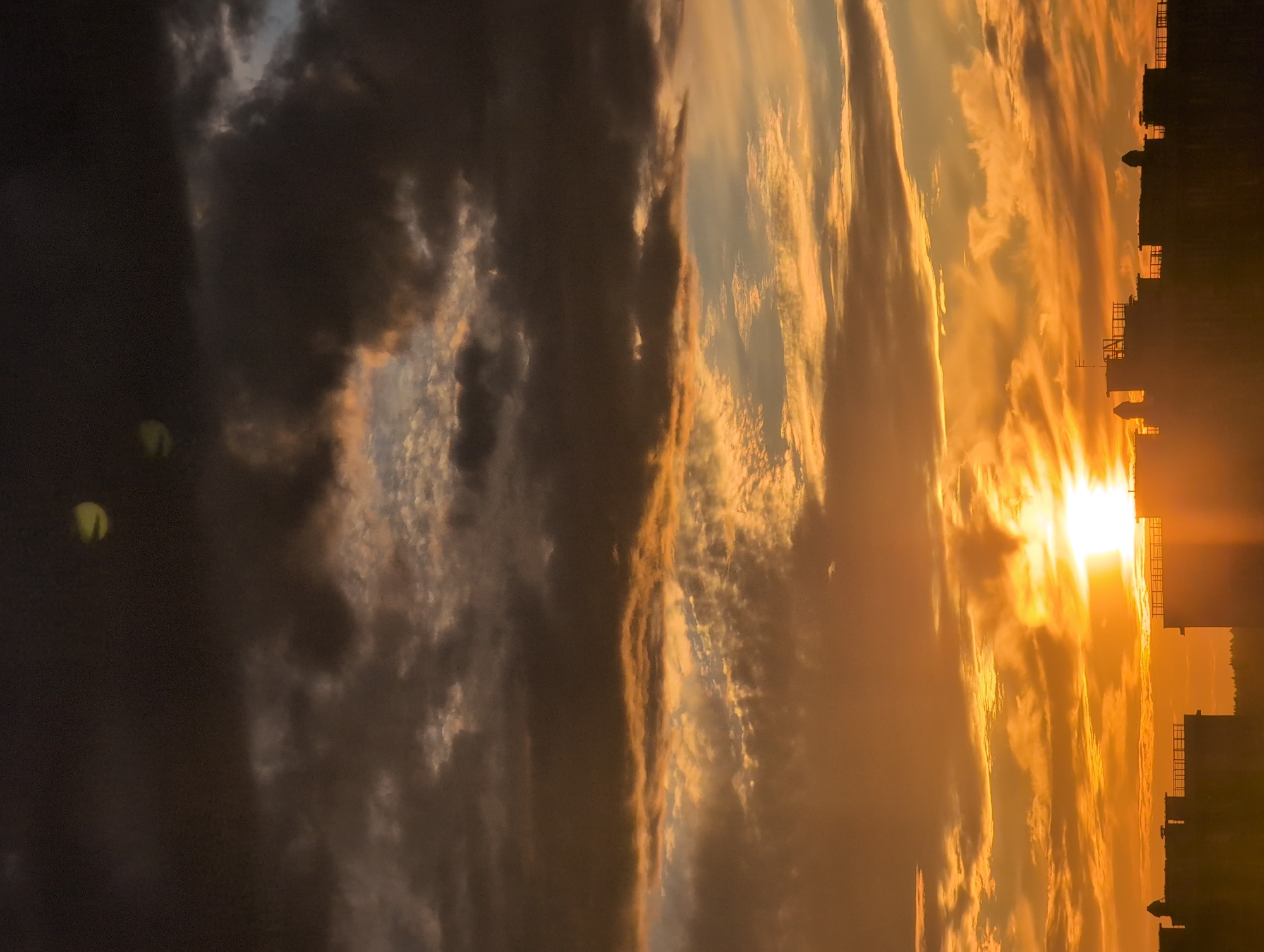 This media file depicts a dramatic sunset over Saint Petersburg, Russia. The sky is filled with a vibrant display of orange and yellow clouds, illuminated by the setting sun. Dark, brooding clouds are interspersed with brighter, wispy formations, creating a sense of depth and movement. In the foreground, the silhouettes of buildings and structures are visible against the fiery sky, with some of them appearing to have intricate architectural details. The sun itself is a bright, intense orb, casting a warm glow across the scene and creating a sense of awe and wonder. The overall impression is one of a breathtaking natural spectacle unfolding over a historic cityscape. The time of day is clearly sunset, characterized by the intense colors of the sky and the long shadows cast by the buildings. The weather appears to be partly cloudy, with a mix of dark and light formations contributing to the dramatic visual effect. The location is identifiable as Saint Petersburg due to the distinctive architectural silhouettes, which are characteristic of the city's skyline. The mood evoked is one of profound beauty and perhaps a touch of melancholy associated with the end of the day. There is no visible text in the media file.