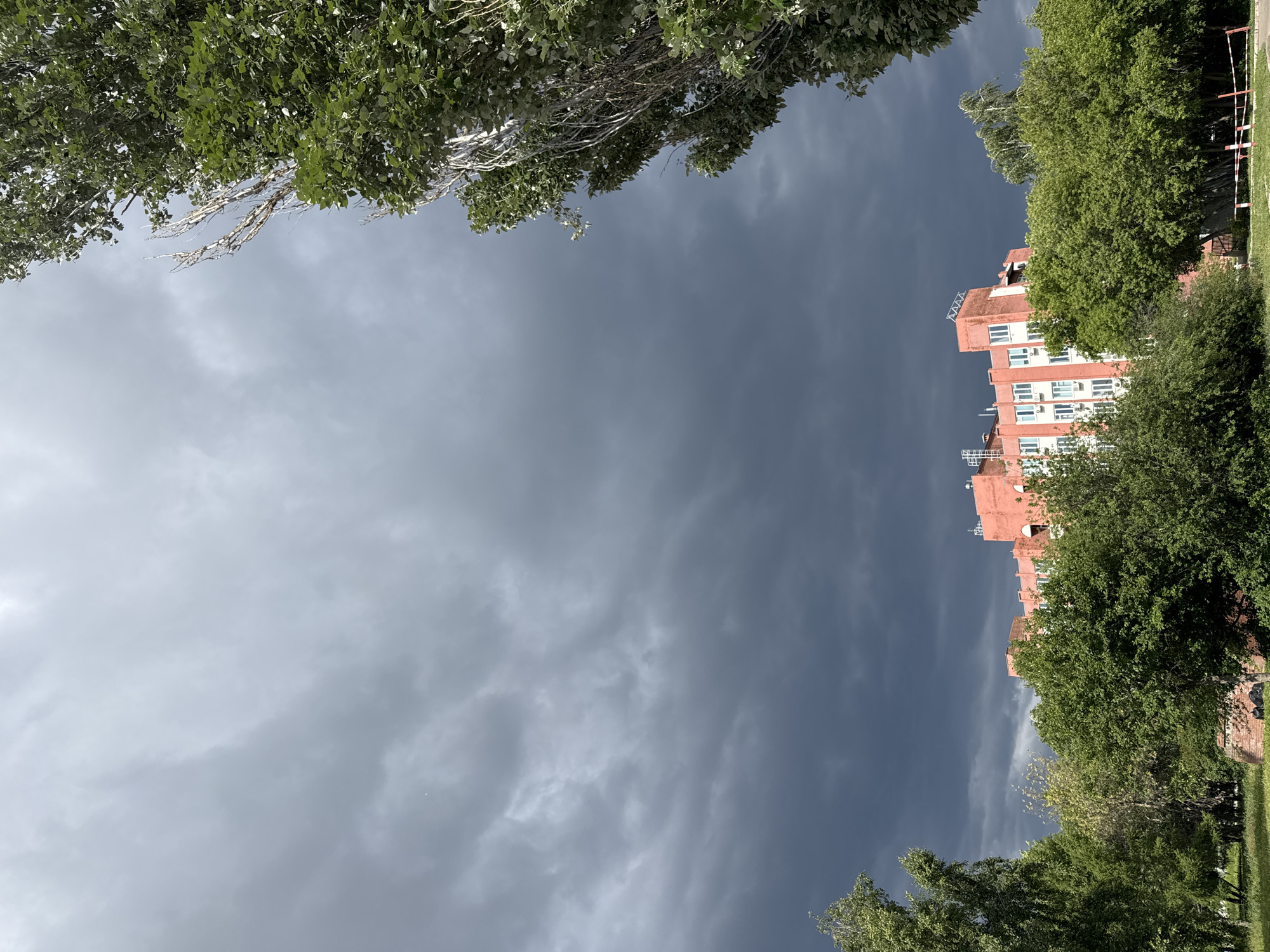 The image depicts an outdoor urban or suburban scene under heavy, dark grey storm clouds, which dominate the upper two-thirds of the frame. In the mid-ground, a multi-story building, painted peach or light orange with white window frames and multiple visible air conditioning units, stands partially obscured by dense green foliage. Architectural elements, possibly ventilation or antenna structures, are present on the building's roof. Tall, leafy trees with bright green foliage flank the scene, notably a large tree on the right with some bare, silvery branches. In the foreground, a band of various green trees and bushes covers the lower portion of the building, with a patch of grass or unkempt ground visible at the bottom. A red and white striped barrier is faintly visible on the lower right, and some dark objects (possibly debris) are partially visible near a low brick structure amidst the foliage in the lower-middle. No individuals or animals are present. The overall lighting is dim, consistent with a heavily overcast sky. The location is in the city of Unknown, Unknown.
