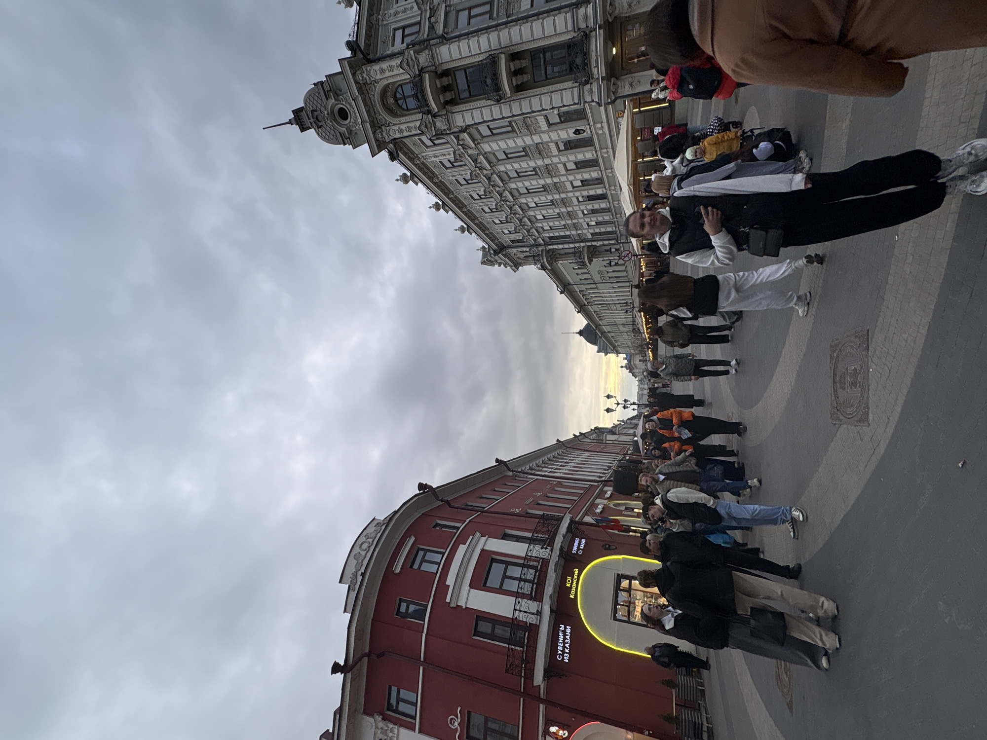 The image depicts a street scene in Kazan, Russia, showcasing architectural elements and a crowd of people. The sky is overcast. On either side of the street, buildings line the road. On the right side is a red building with visible windows, balconies, and a curved facade. Illuminated signage with Cyrillic text is visible on the building. On the left side, a lighter colored, ornate building with detailed window structures and architectural embellishments stands tall. A crowd of people is walking on the paved street, with individuals wearing a variety of clothing. The street is patterned with brick and stone.