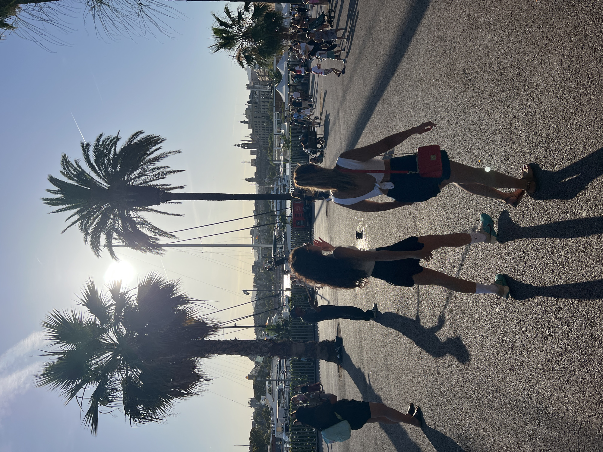 This image captures a vibrant, sun-drenched scene on a pedestrian promenade adjacent to a marina in Barcelona, Spain, specifically appearing to be Port Vell. The late afternoon sun, low and positioned centrally between two large palm trees on the left, creates strong backlighting, producing long, defined shadows on the dark asphalt pavement and a prominent lens flare. Contrails streak across the clear blue sky.
In the foreground, two women walk away from the camera. The woman on the right, with blonde hair, wears a white crop top tied at the back, black shorts, and brown flat sandals, carrying a small red shoulder bag. To her left, another woman with long, dark curly hair wears a light-colored top, black shorts, white socks, and light blue athletic shoes, with her right hand raised as if in a wave or gesture.
Further into the midground, a third woman on the far left wears black clothing and a light blue backpack, holding a mobile phone. A man in dark attire stands beneath the central palm tree, appearing to interact with something off-camera. The promenade is populated by numerous other people, including families with children and individuals strolling, some gathered near white tent structures.
The background showcases a marina densely packed with various yachts and sailboats, separated from the promenade by a low fence. Beyond the marina, the distinctive skyline of Barcelona's city center rises, featuring historic buildings with spires and characteristic European architecture bathed in the golden light of the setting sun.