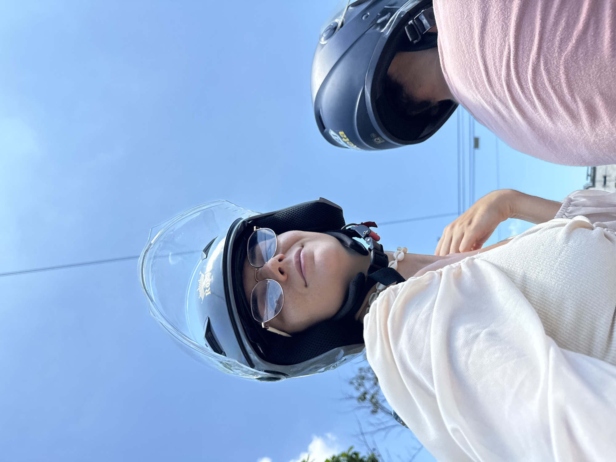 Two individuals wearing helmets are on a scooter, with a bright blue sky and some thin power lines visible in the background. The person in the foreground, positioned on the left, is a woman wearing round, gold-rimmed glasses and a white, flowy top. She is looking slightly upwards and to the right. A seashell necklace is visible around her neck. Behind her, to the right, is another person wearing a dark helmet and a pink t-shirt. The scene appears to be outdoors, possibly in a tropical or subtropical urban environment, given the clear sky and the presence of a scooter, which are common modes of transportation in cities like Hồng Sơn, Vietnam.