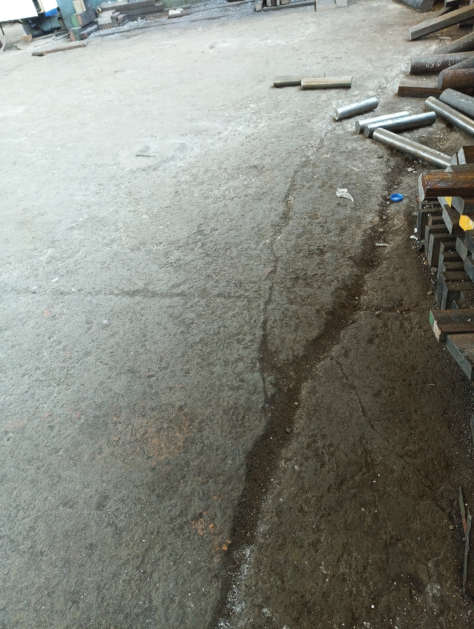 The image depicts an outdoor, possibly industrial setting. A concrete floor, appearing somewhat worn and damp in areas, occupies the majority of the frame. Various metal rods and beams, some cylindrical and others rectangular, are scattered and stacked on the floor. A small blue cap or lid is visible on the ground. In the background, parts of machinery or equipment are faintly visible, along with additional stacked materials. The scene suggests a storage or work area within a facility.