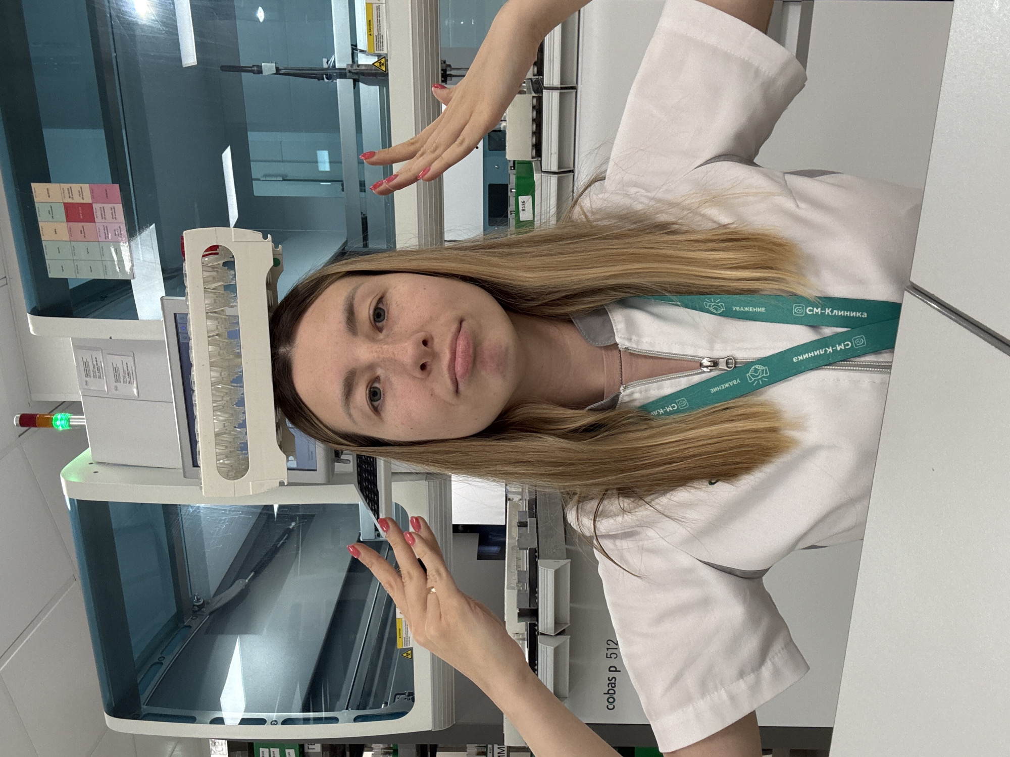 A young Caucasian woman with blonde hair is standing in what appears to be a medical laboratory. She is wearing a white lab coat with a green lanyard around her neck, which has Cyrillic text and logos visible. On top of her head, she has balanced a rack of small vials. Her hands are raised on either side of her head, with fingers splayed, and she is looking at the camera with a slightly pursed expression. In the background, there is a large piece of laboratory equipment, possibly a diagnostic machine, with a blue transparent cover and a control panel. Text indicating "cobas p 512" is visible on the equipment. The environment suggests a sterile, professional setting. The image does not provide specific details about the location within Moscow, Russia.