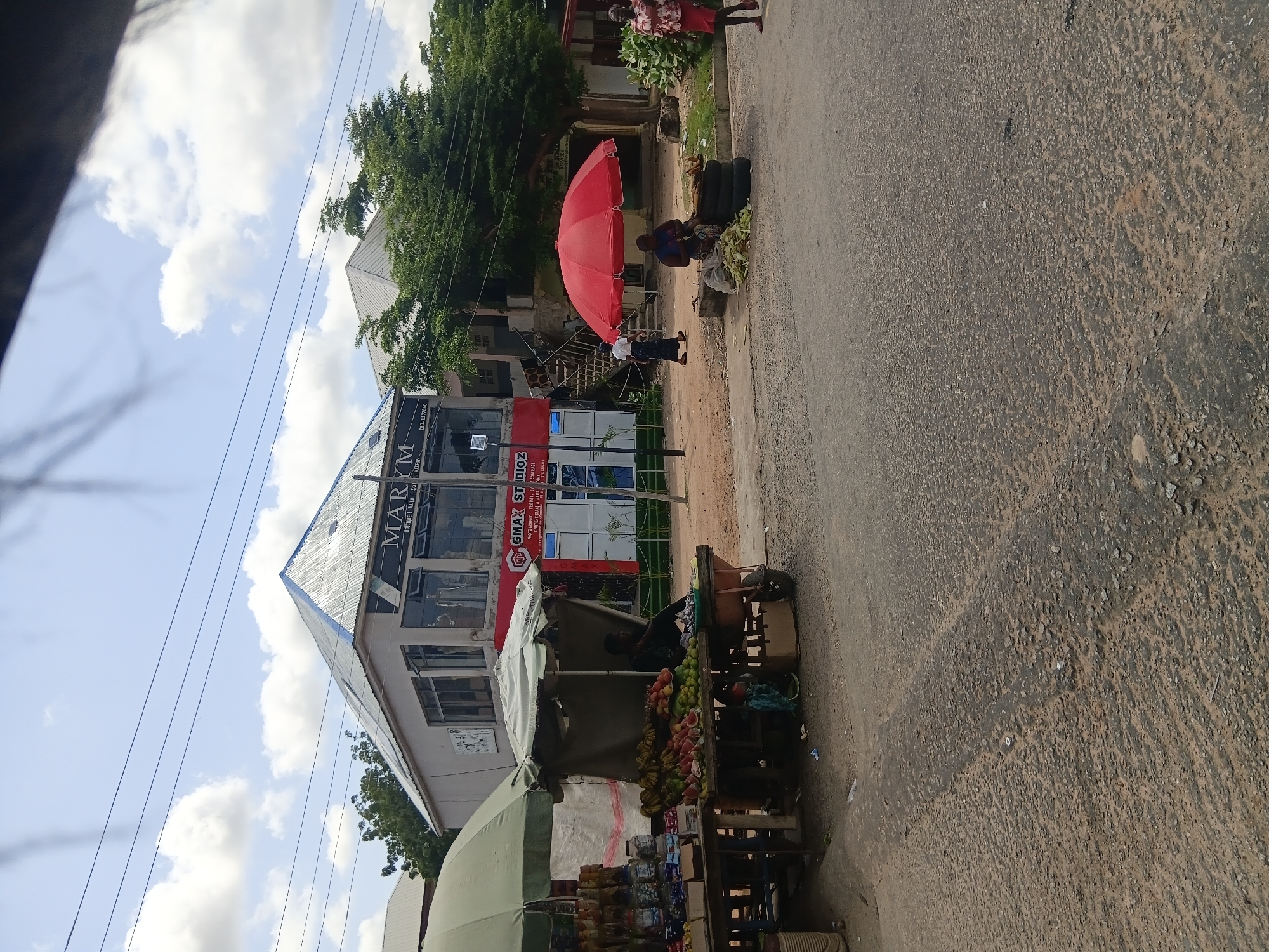 The image depicts a street scene in Jalingo, Nigeria. A building with a prominent sign reading "MARYM BOUTIQUE | NAILS | MAKEUP" is visible in the background. In front of the building, a busy street market is set up. Several stalls display an assortment of fruits, including watermelons, bananas, and other produce. One stall is covered by a large red umbrella, under which two individuals are seated, possibly selling goods. Another stall, covered by a green umbrella, displays bottles and packaged items. Pedestrians are seen walking along the street, and a person is seen walking towards the market stalls. The environment appears to be a typical urban street with a mix of commercial buildings and market activity. The road surface is paved with asphalt and shows signs of wear. The sky is partly cloudy.