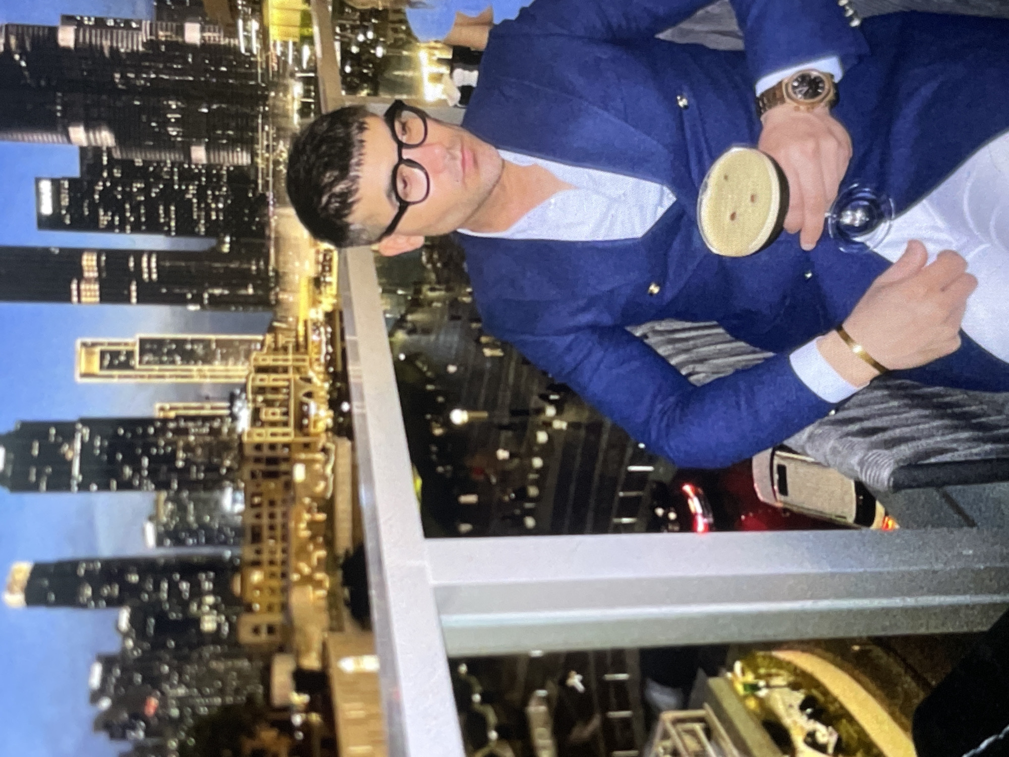 An overhead medium shot shows a man in a blue suit sitting at a table on a balcony overlooking a city skyline at dusk. He is wearing glasses and holding a cocktail. The city skyline features numerous tall buildings lit up with lights. The man's hands are visible, one holding the cocktail glass and the other resting on the table. The balcony railing is in the foreground.