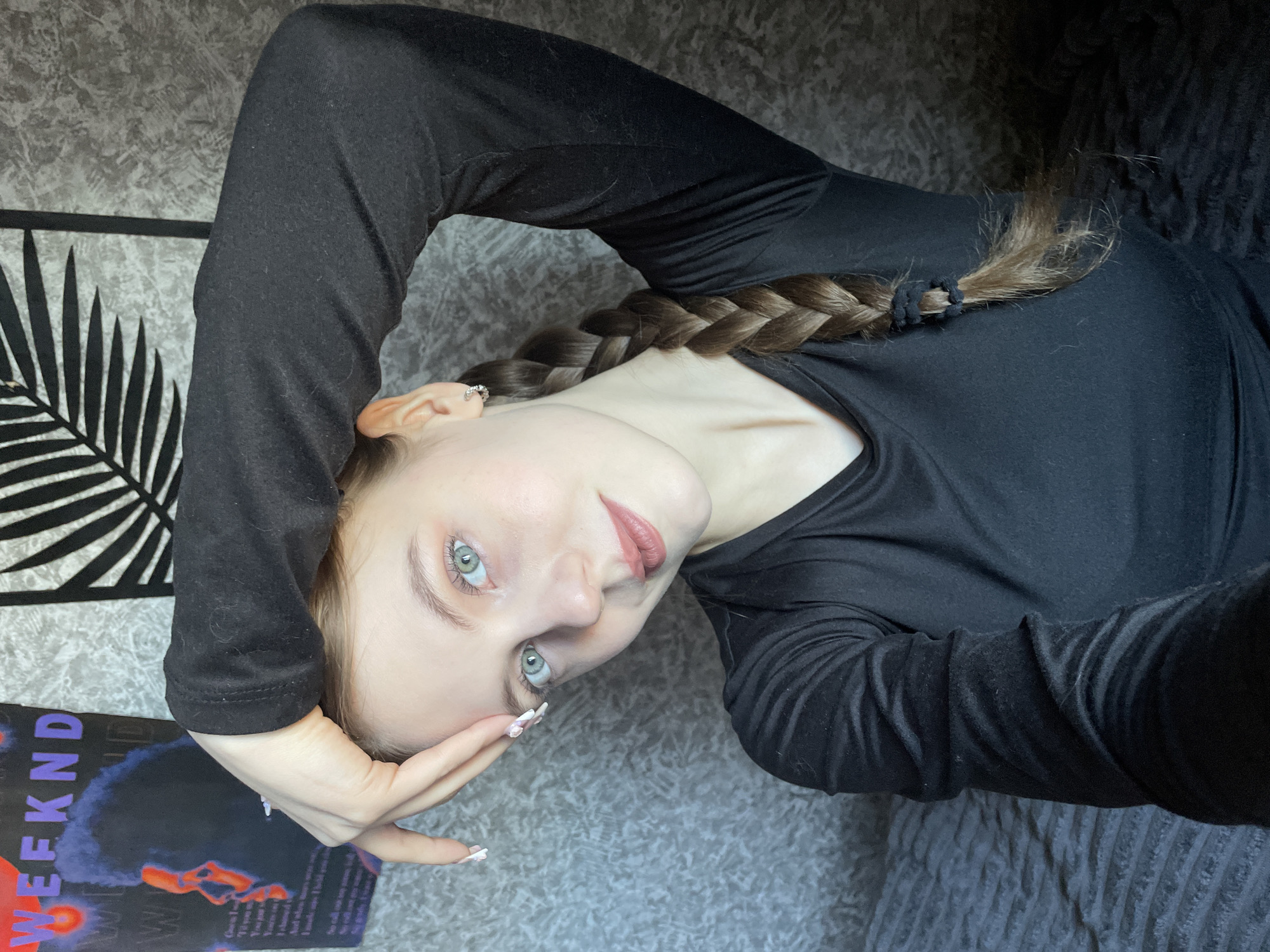 A fair-skinned young woman with blue-green eyes and a brown braided hairstyle is lying on what appears to be a gray textured sofa. She is wearing a long-sleeved black top. Her head is tilted to her right and is being supported by her right hand. Her left arm extends over her head towards a black and white framed artwork depicting palm leaves. A magazine with the word "WEEKND" is visible next to the artwork. The location cannot be verified as Фоки, Russia based on the provided image.