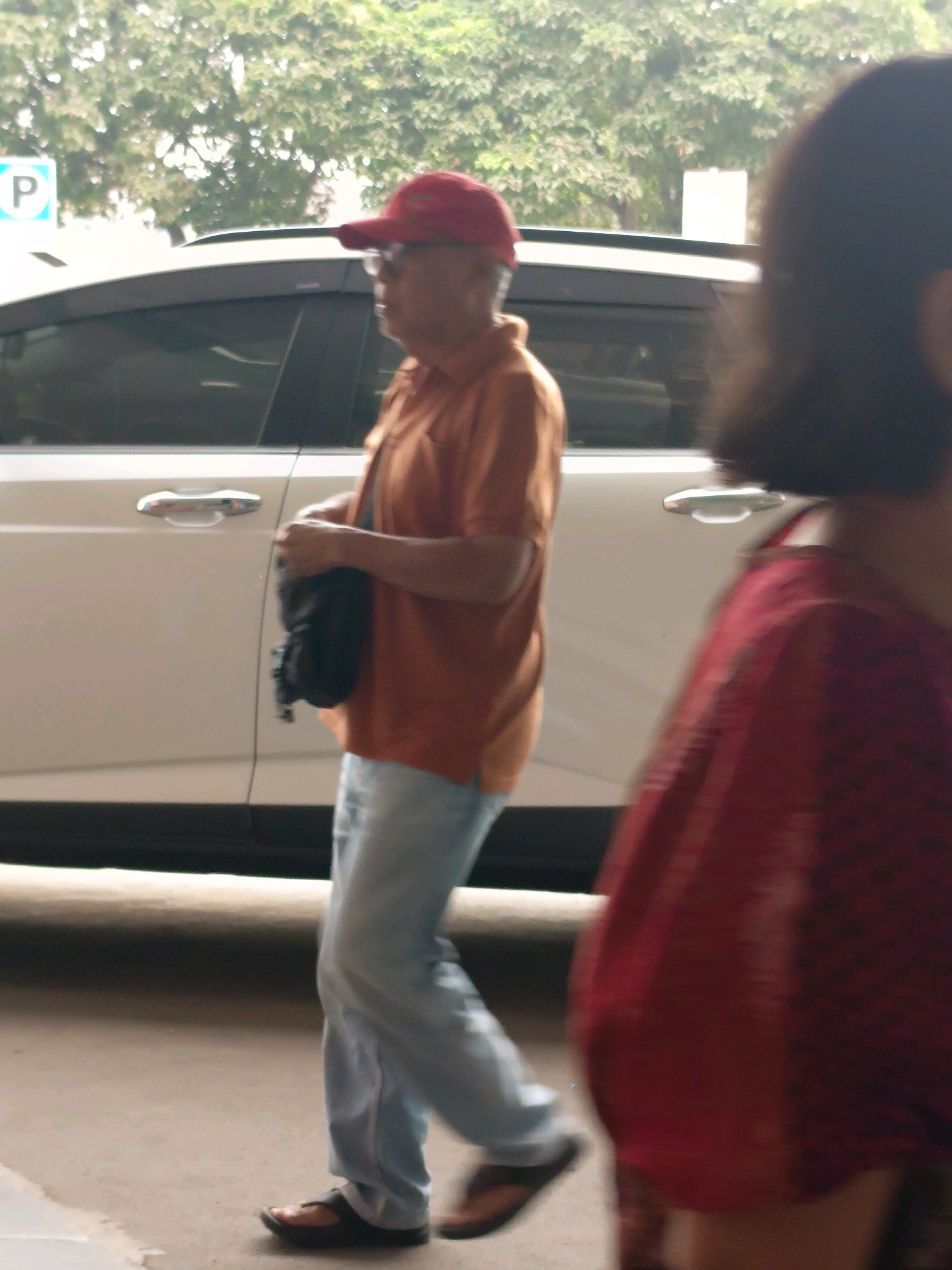 The image captures a street scene in Tangerang, Indonesia, with a man in the foreground and a blurred figure of a woman to the right. The man, wearing a red cap, glasses, and an orange collared shirt, is walking to the left, carrying a black bag over his left shoulder. He is wearing light blue jeans and dark sandals. Behind him is a white vehicle with chrome door handles. The background is dominated by foliage from trees and what appears to be a parking sign with a blue circle and a white "P". The ground is paved with concrete. The woman to the right is mostly out of focus and is wearing a red garment.