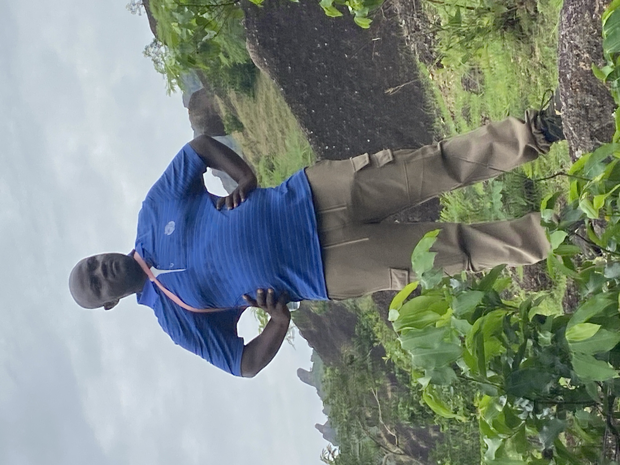 A man is standing outdoors in what appears to be a hilly, rocky landscape in Ganawuri, Nigeria. He is wearing a blue striped polo shirt, khaki pants, and sneakers. He has his hands on his hips, and he's looking toward the camera. The environment consists of rocks, vegetation, and a cloudy sky.