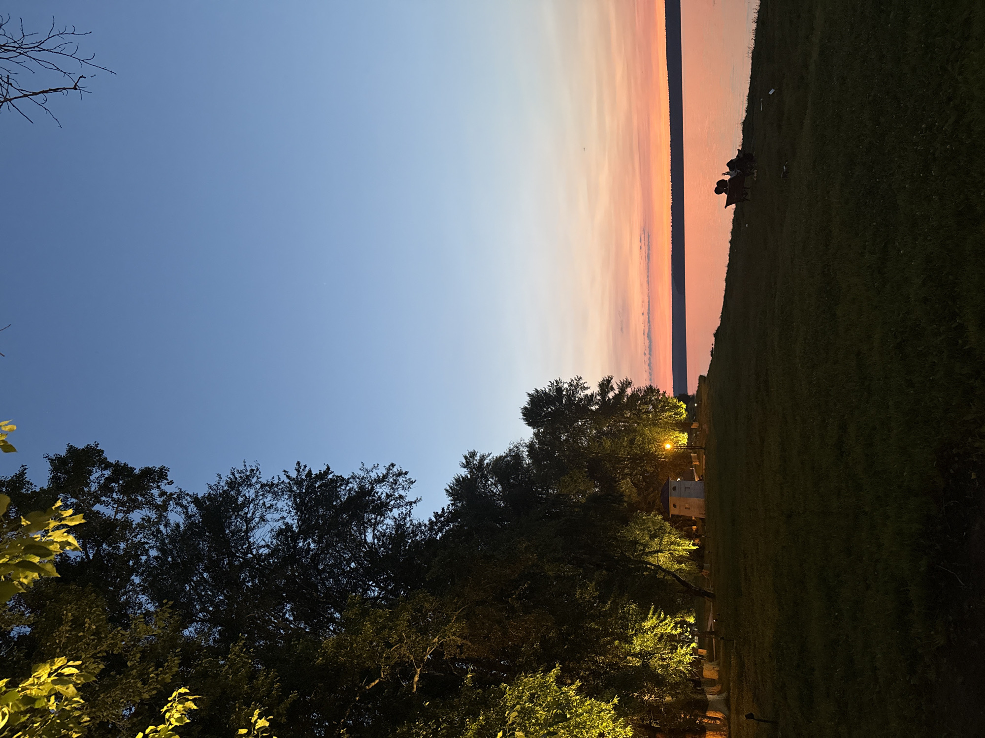 The image depicts a serene evening scene along the shore of Lake Votkinsk in the city of Votkinsk, Russia. On the left, a line of trees is illuminated by artificial lights, casting a warm yellow glow on their foliage and the grassy embankment below. Beyond the trees, a small, light-colored, square building with a dark roof and a single window is visible. A lamppost stands nearby, also illuminated. In the foreground, a well-maintained grassy area slopes down towards the water.
On the right side of the image, two figures are seated in chairs facing the lake, their silhouettes clearly defined against the water. They appear to be relaxed, possibly enjoying the sunset. The lake itself is calm, reflecting the soft hues of the twilight sky, which transitions from a deep blue at the top to shades of pink and orange near the horizon. A thin, dark line of trees marks the distant shore across the water. The overall atmosphere is peaceful and tranquil, suggesting a quiet evening by the lake.