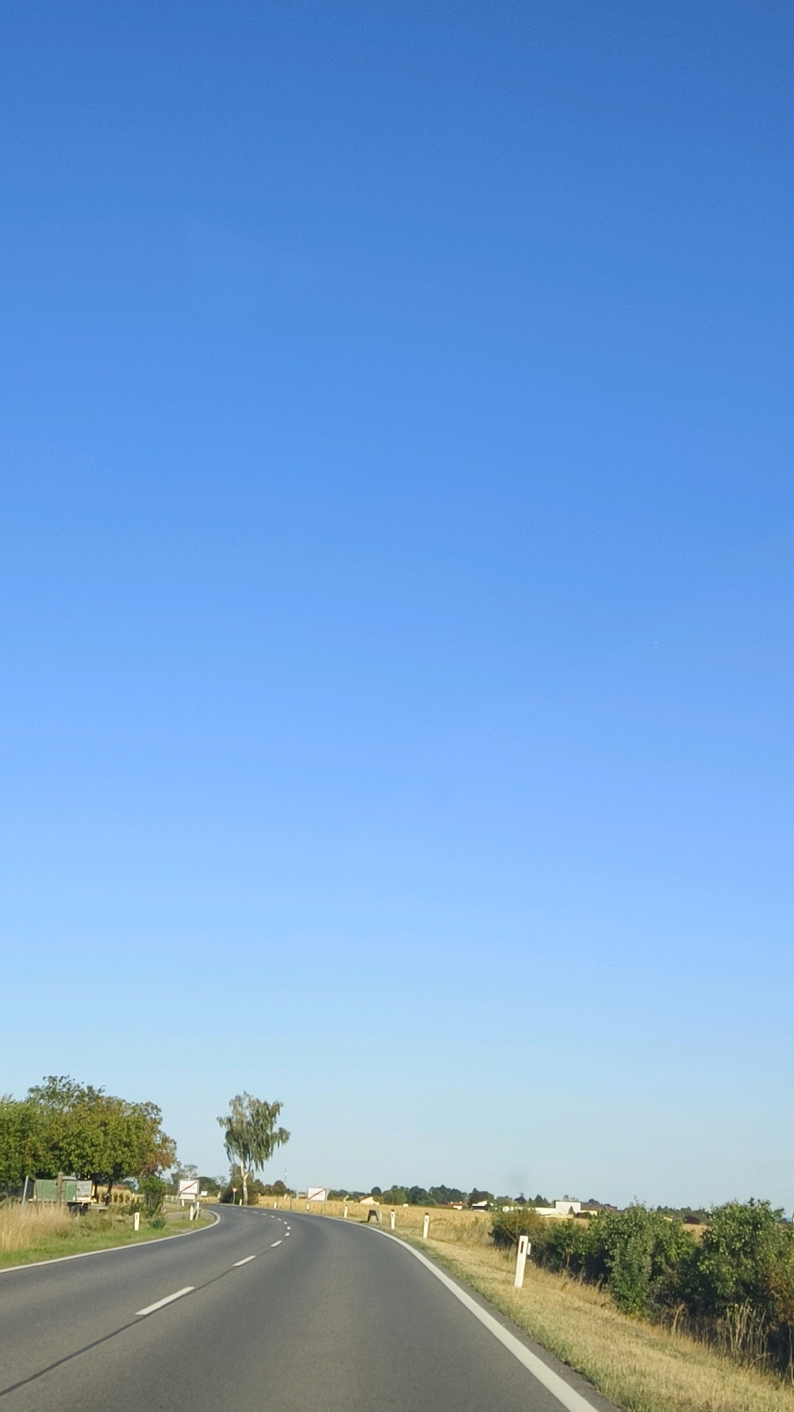 The image shows a paved road curving to the right through a rural landscape under a clear blue sky. The road has white dashed and solid lines indicating lanes. On the left side of the road, there are trees and a structure that appears to be a truck. On the right side of the road, there is dry grass, bushes, and white posts marking the roadside. In the distance, there are more trees and buildings, suggesting a small settlement. The scene is likely captured in the city of Feuersbrunn, Austria, given the characteristic rural setting with agricultural land and roadside markers common in the region. There are no people or animals visible in the image. The overall impression is one of a serene and open countryside, possibly during daytime or late afternoon given the clear sky and lighting.