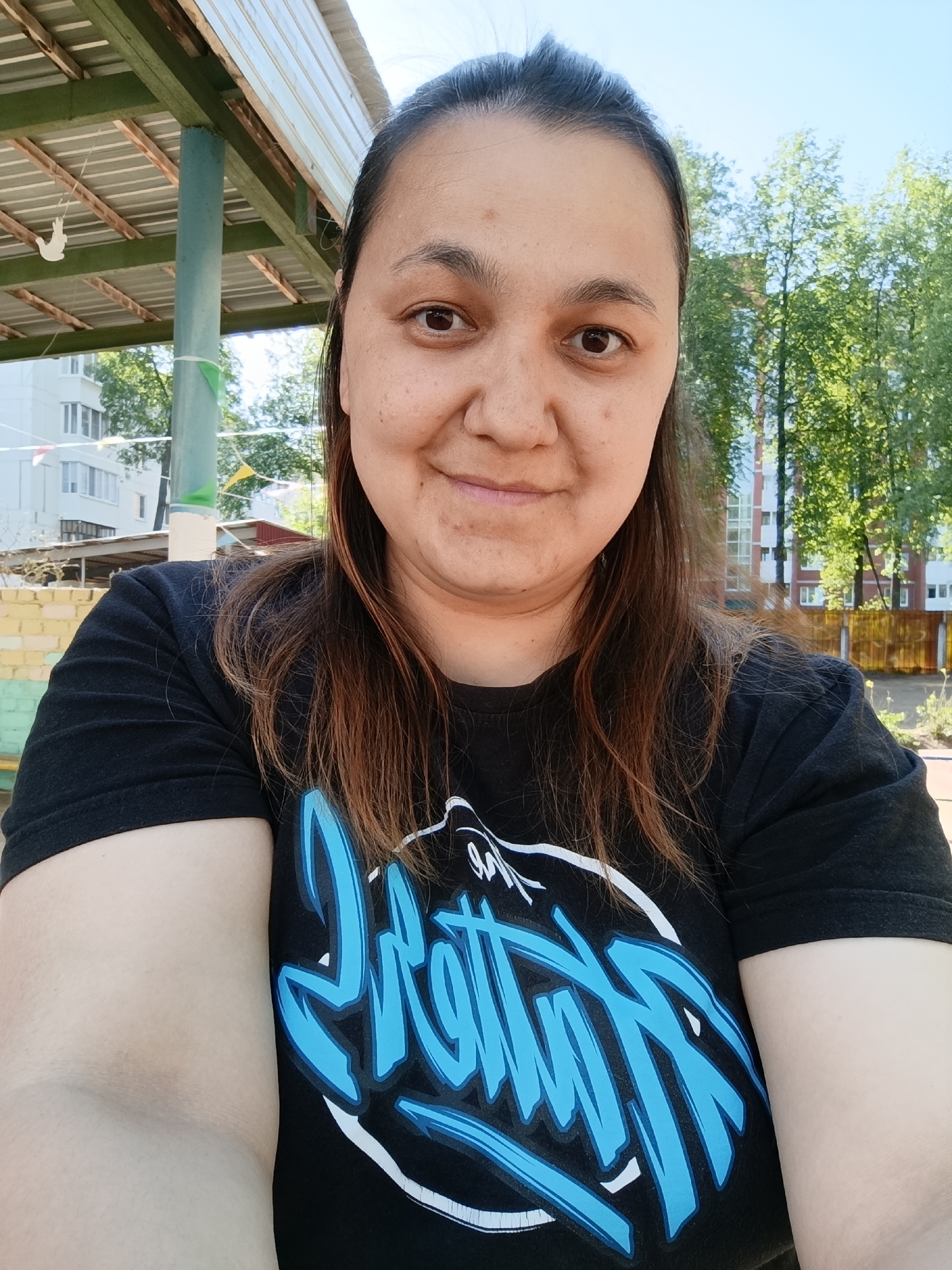 A woman is taking a selfie outdoors. She is wearing a black t-shirt with a blue and white graphic. She has dark hair pulled back and a slight smile.
Behind her, there is a covered structure with hanging decorations. Buildings and trees are visible in the background. Based on the information provided, the location is Chaikovsky, Russia.