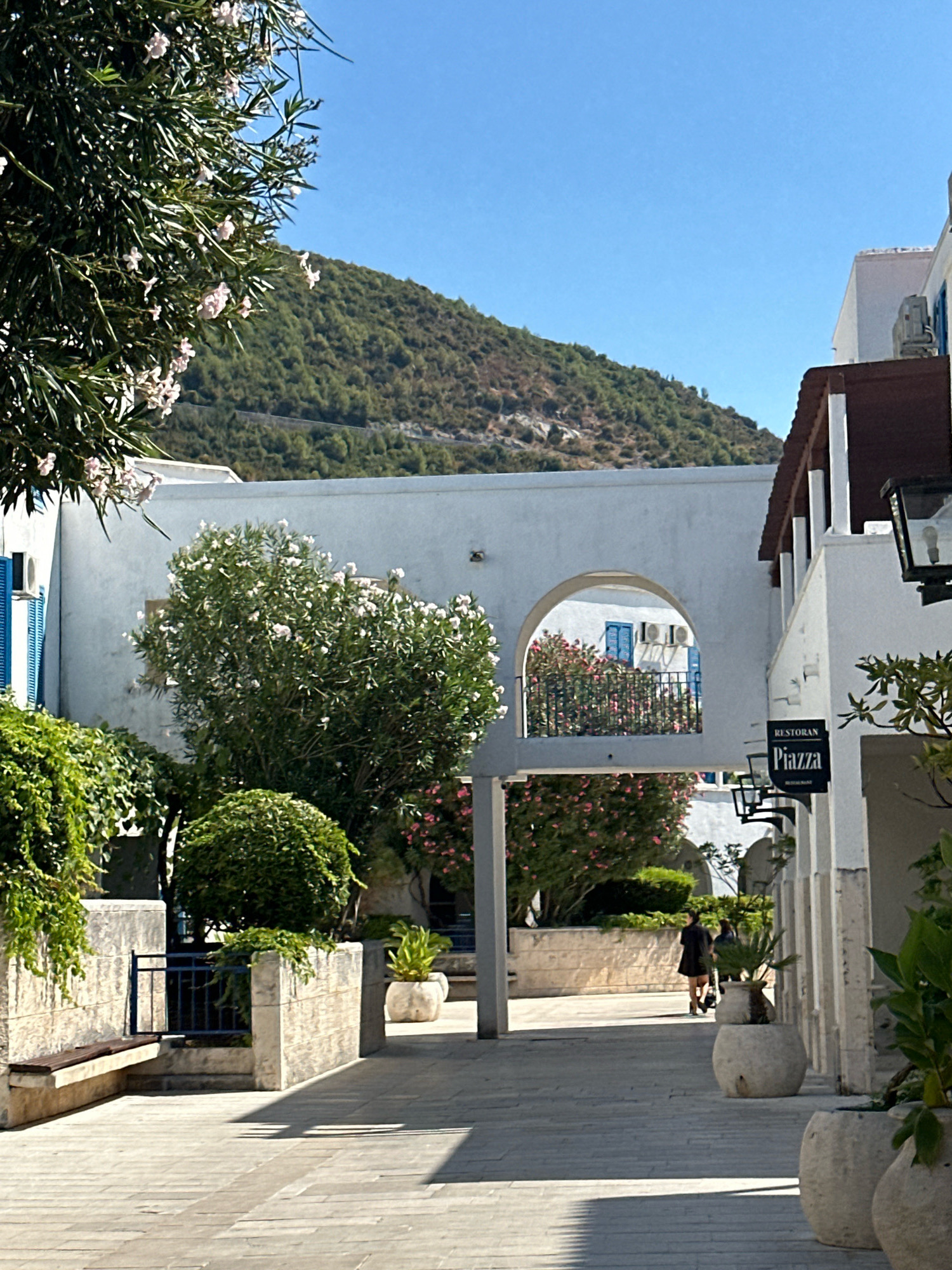 The image shows a narrow paved pathway in a Mediterranean coastal town, likely Budva, Montenegro, on a sunny day. The pathway is flanked by white-washed buildings and lush greenery. On the left, a bench is set against a stone wall draped with ivy. A dark blue metal fence separates the bench area from a planted area with a rounded, manicured bush. Ahead, an arched walkway connects two buildings, revealing a glimpse of a balcony with pink flowers and blue shutters. On the right, a building features a dark brown awning supported by white columns, and a black sign with white lettering reads "RESTORAN Piazza". A woman in a dark dress is walking away from the camera down the pathway, and another figure is partially visible behind her. The background features a sloping, tree-covered hillside under a clear blue sky. Large potted plants are visible along the pathway.