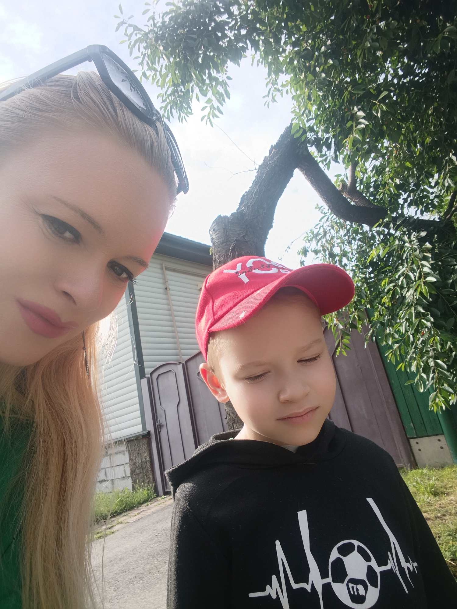 In a selfie taken outdoors, a woman with blonde hair and sunglasses on her head is positioned in the foreground on the left, with a young boy wearing a red baseball cap and a black hoodie in the foreground on the right. The boy is looking down with his eyes closed. Behind them, a tree with green leaves is visible, along with a building with horizontal siding and a dark door. The overall setting appears to be a residential area, possibly a backyard or a street in the city of Chernihiv, Ukraine. The lighting suggests it is daytime.