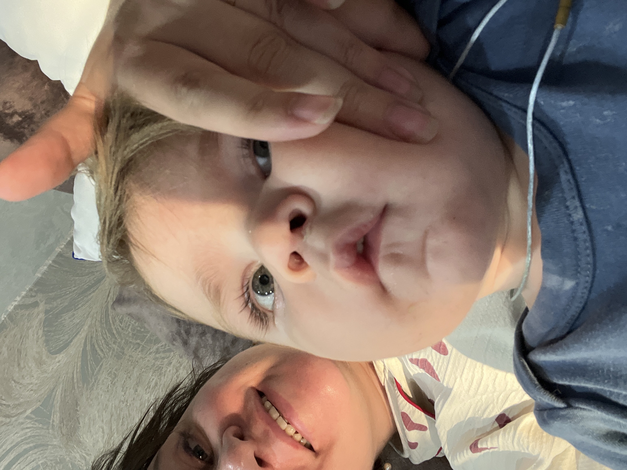 The image is a close-up selfie of a woman and a baby. The woman is on the left, smiling, and her face is partially obscured by the baby. She has dark hair and is wearing a white shirt with a pattern. The baby is in the center and right of the image, looking directly at the camera. The baby has fair hair and is wearing a blue shirt. A person's hand is touching the baby's face. The background appears to be a bed with a patterned duvet and a pillow. The scene is indoors. There is no information available to determine the specific location in the city of Noviy, Russia.