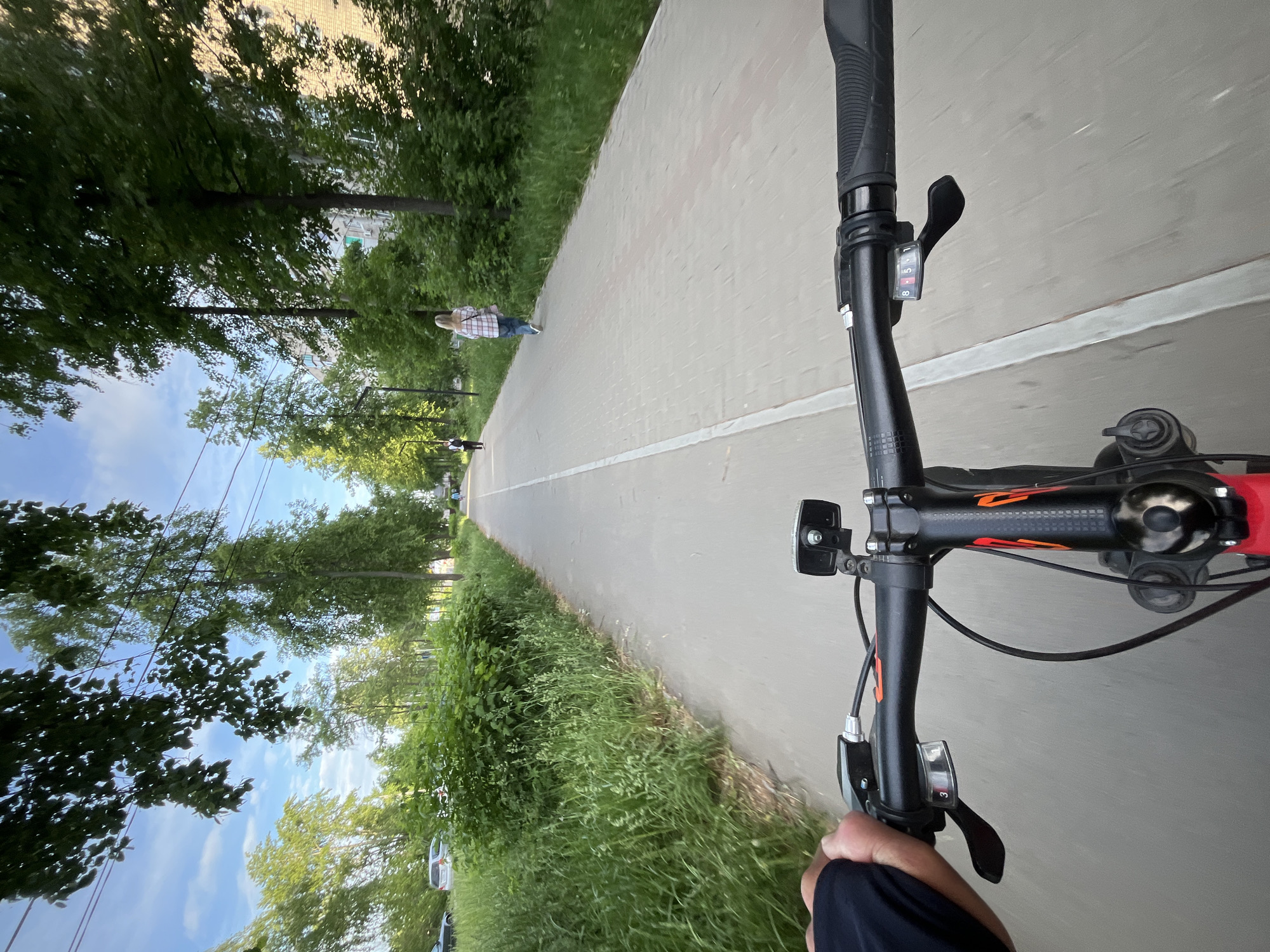 View from a bicycle's perspective on a paved path in Chaikovsky, Russia. The path is bordered by green vegetation on both sides, with trees lining one edge. A white line marks the center of the path. Two pedestrians are visible ahead on the path, one wearing a plaid shirt. The bicycle handlebars are in the foreground, showing the gear shifter, light, and other controls. A hand holding the handlebar is visible on the right side. The sky is visible between the trees, with buildings in the background. The road is paved with interlocking concrete pavers.