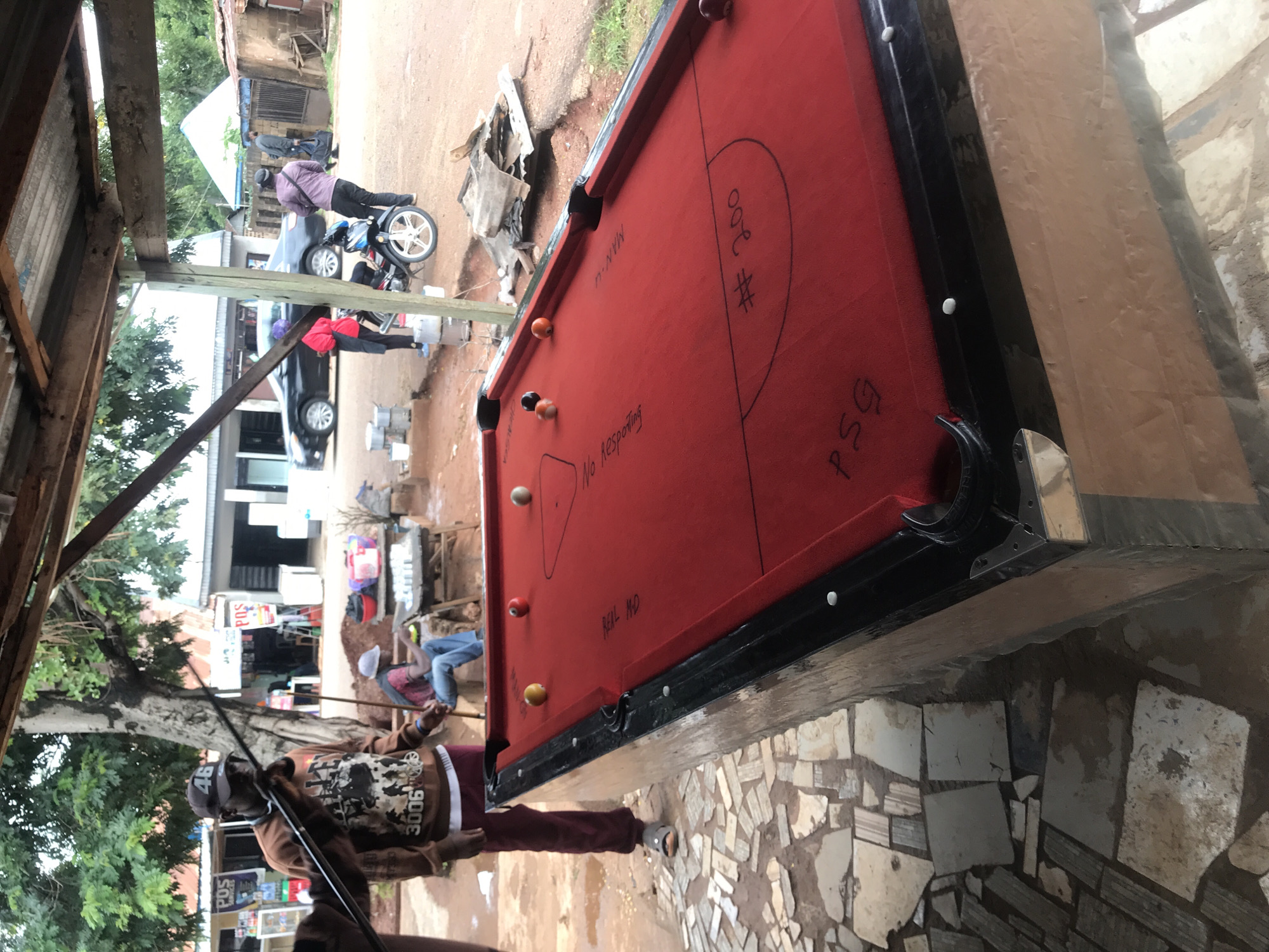 This media file depicts an outdoor scene in Kabong, Nigeria, likely in the afternoon. The main focus is a red pool table situated on a paved area. Several people are visible in the background, engaged in various activities.
In the foreground, a man wearing a brown hooded sweatshirt with "46" and "3006" visible on it, and a dark cap, is holding a pool cue, suggesting he is either playing or about to play pool. To his right, another person is seated on what appears to be a bench. Further in the background, several individuals are standing or walking near a motorcycle and a dark-colored car. The environment appears to be somewhat informal, with visible structures like a wooden shelter on the left and buildings with corrugated iron roofs in the distance. The ground is a mix of dirt and stone paving.
The pool table itself has some markings on its surface, including what appears to be "MAN U", "# 200", "No Responding", and "PSG". There are also several pool balls scattered on the table, including an orange ball and a brown ball.
The overall atmosphere seems casual and everyday. The lighting suggests it is daytime, and the presence of trees indicates a relatively green environment, though the ground appears dry. The scene captures a moment of leisure and social interaction in a Nigerian community.