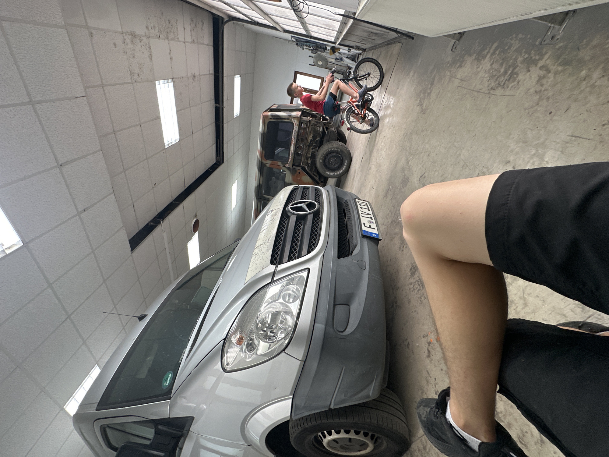 The image shows a young boy balancing on a bicycle in a garage. He is wearing a red t-shirt and blue shorts. The bicycle is an orange BMX bike. Behind the boy is a derelict vehicle that appears to be a heavily modified van, possibly a former Mercedes-Benz Sprinter, with rust and exposed metal. The garage has a white tiled ceiling with fluorescent lights and a large open door at the far end, revealing a bright, possibly sunny, outdoor environment. The floor is concrete.
In the foreground, a person's legs are visible, wearing black shorts and black sneakers with white socks, suggesting they are sitting or lying down and filming or photographing the scene. The overall impression is a casual, perhaps playful, moment captured in a utilitarian setting. The location is identified as Mainz, Germany. No specific time of day or weather conditions are explicitly evident, other than the brightness from the open garage door implying daylight. The mood appears to be relaxed and informal. The license plate on the derelict vehicle reads "F LV 117".