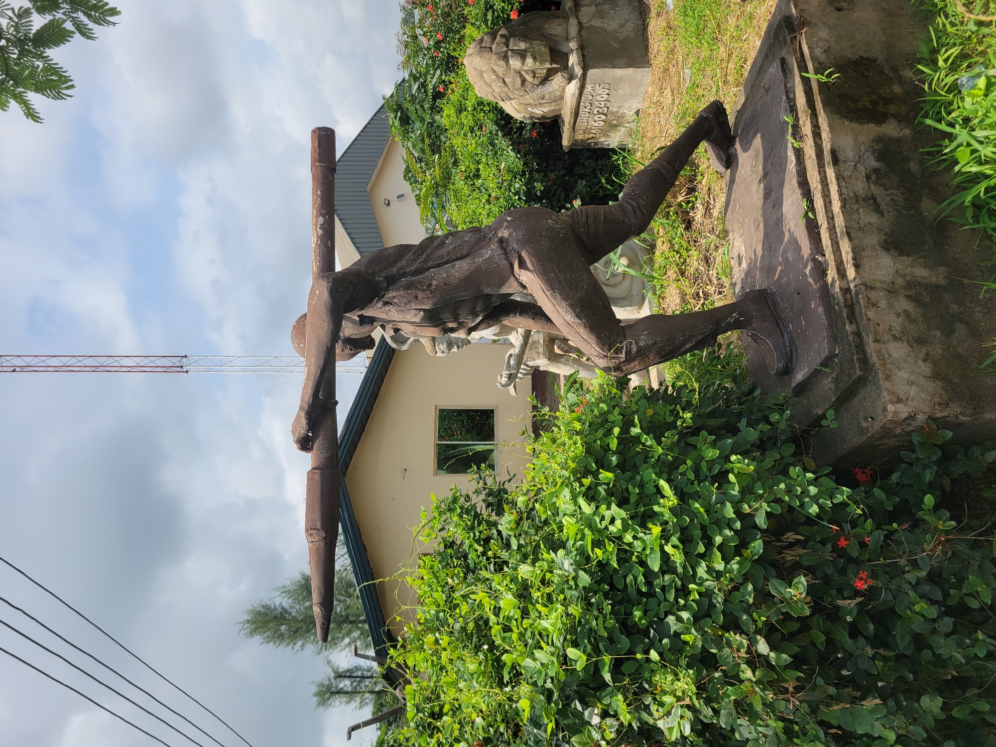 The image depicts a statue of a man in a dynamic pose, with his arms extended and holding a horizontal cylindrical object across his shoulders. The statue is made of a dark, textured material, possibly bronze or concrete, and shows signs of weathering. It is situated outdoors on a concrete base, surrounded by lush green vegetation. In the background, a light-colored building with a dark-sloped roof is visible, along with a tall radio tower. To the right of the main statue, a stone sculpture of a lion's head is mounted on a pedestal. The overall environment suggests an outdoor exhibition or public space. The image does not provide explicit visual clues to definitively place it within Egbagu, Nigeria, but the description implies such a context.