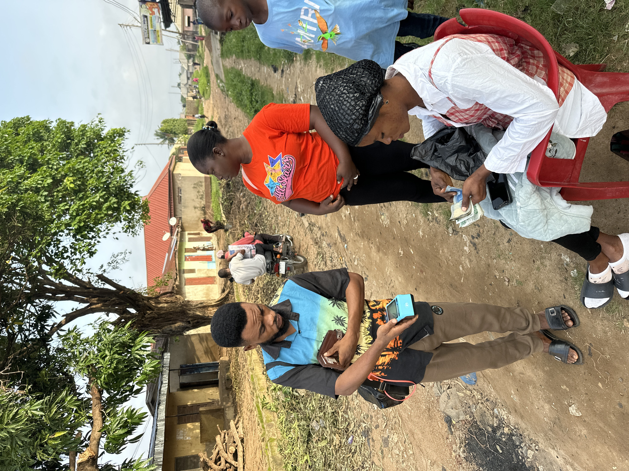 In Narayi, Nigeria, a man in a tropical print shirt and brown trousers stands on a dirt road, holding a brown wallet and a blue card reader. Beside him, a woman in an orange shirt and black pants looks to her left. In front of them, a woman with a black hair net sits in a red chair, holding cash and a black plastic bag, with a mobile phone on her lap. To the right, a young boy in a blue shirt with a bird graphic stands and looks to the left. The background shows several buildings with red roofs, a large tree, and power lines, suggesting a semi-urban or rural setting. Vegetation and debris are scattered on the ground.