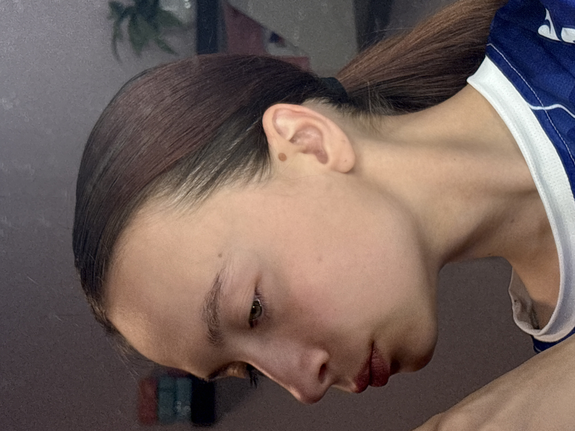 A close-up, indoor photograph shows a person's face in profile. The individual has brown hair pulled back in a ponytail, light skin, and visible facial features including an eye, nose, and mouth with dark lipstick. A mole is visible near the ear. The person is wearing a blue and white athletic-style shirt. The background is blurry and indistinct, suggesting an interior space. There is no information about the location being in Прикамский, Russia.