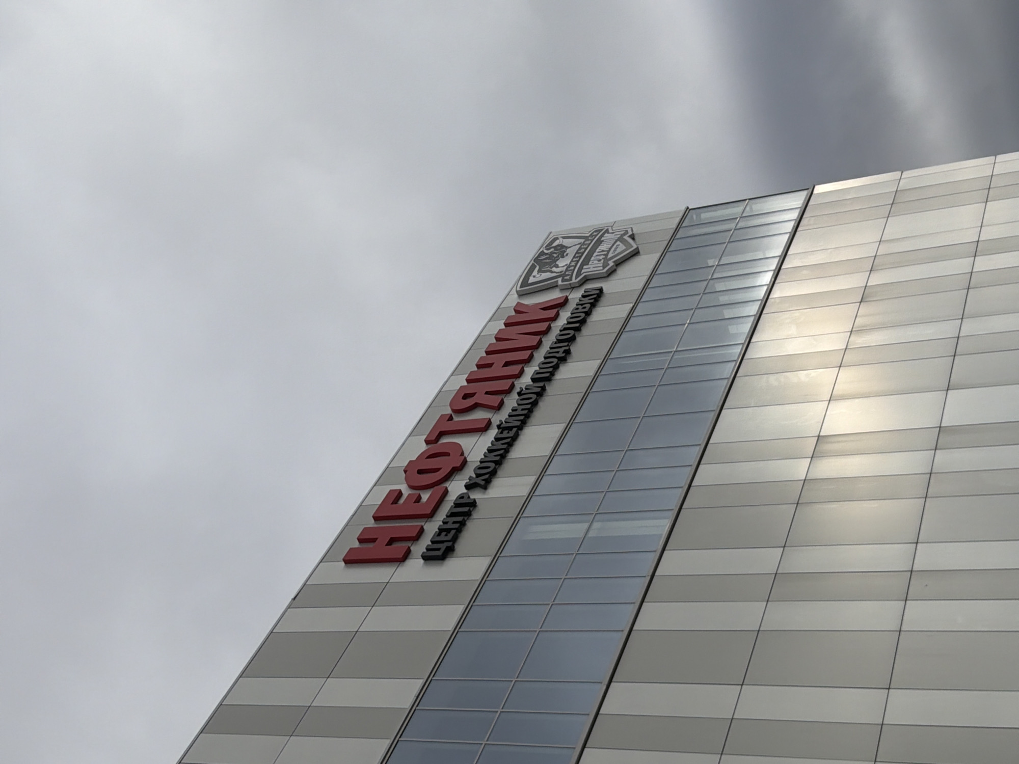 A low-angle shot depicts the facade of a building in Almetyevsk, Russia. The building's exterior is constructed from horizontally arranged, alternating shades of gray panels. A vertical section of glass windows runs along the side of the building. In red and black Cyrillic script, the name "NEFTEKHIMIK" is displayed prominently, indicating the building likely belongs to or represents the Neftekhimik hockey organization, which is based in Almetyevsk, Russia. A crest is above the name. The sky above is overcast and gray.