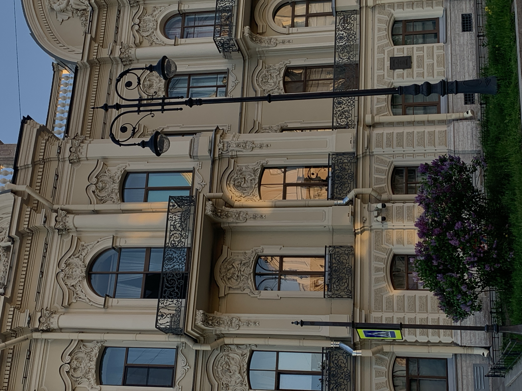 The image depicts a section of an ornate, multi-story building in Saint Petersburg, Russia. The facade features arched windows with decorative surrounds, wrought-iron balconies, and elaborate ornamentation. A vintage street lamp with two globe lights extends horizontally across the frame. To the right, a flowering lilac bush is visible. A crosswalk sign is attached to a light post. The building appears to be well-maintained and reflects the historical architectural style of the city. The lighting suggests either dawn or dusk. There are no people visible in the image.