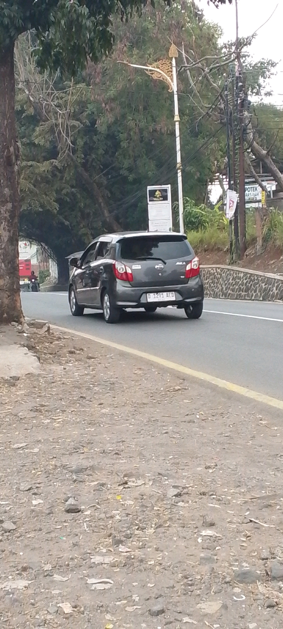 A dark gray hatchback is driving on a paved road in Cilaku, Indonesia. The car is facing away from the camera, and its license plate reads "D 1291 AED". The road is bordered by a dirt shoulder on the left and a stone wall on the right. Trees and vegetation line the road, and a street light is visible in the background. There are also power lines and some signs on poles. The overall scene depicts a typical roadside environment in a moderately vegetated area.