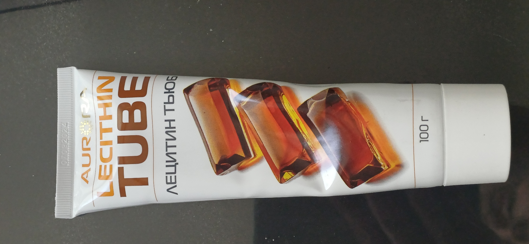 A white tube of "LECITHIN TUBE" with Russian text "ЛЕЦИТИН ТЮБ" and an image of amber-colored capsules is presented on a dark, textured surface. The tube has gold lettering for the brand name "AURORA" and the product name. At the top of the tube, the date "07092024" is printed. Near the bottom, "100 г" (100 grams) is indicated. The background is dark and appears to be a countertop or table. There are no people or discernible city locations visible.