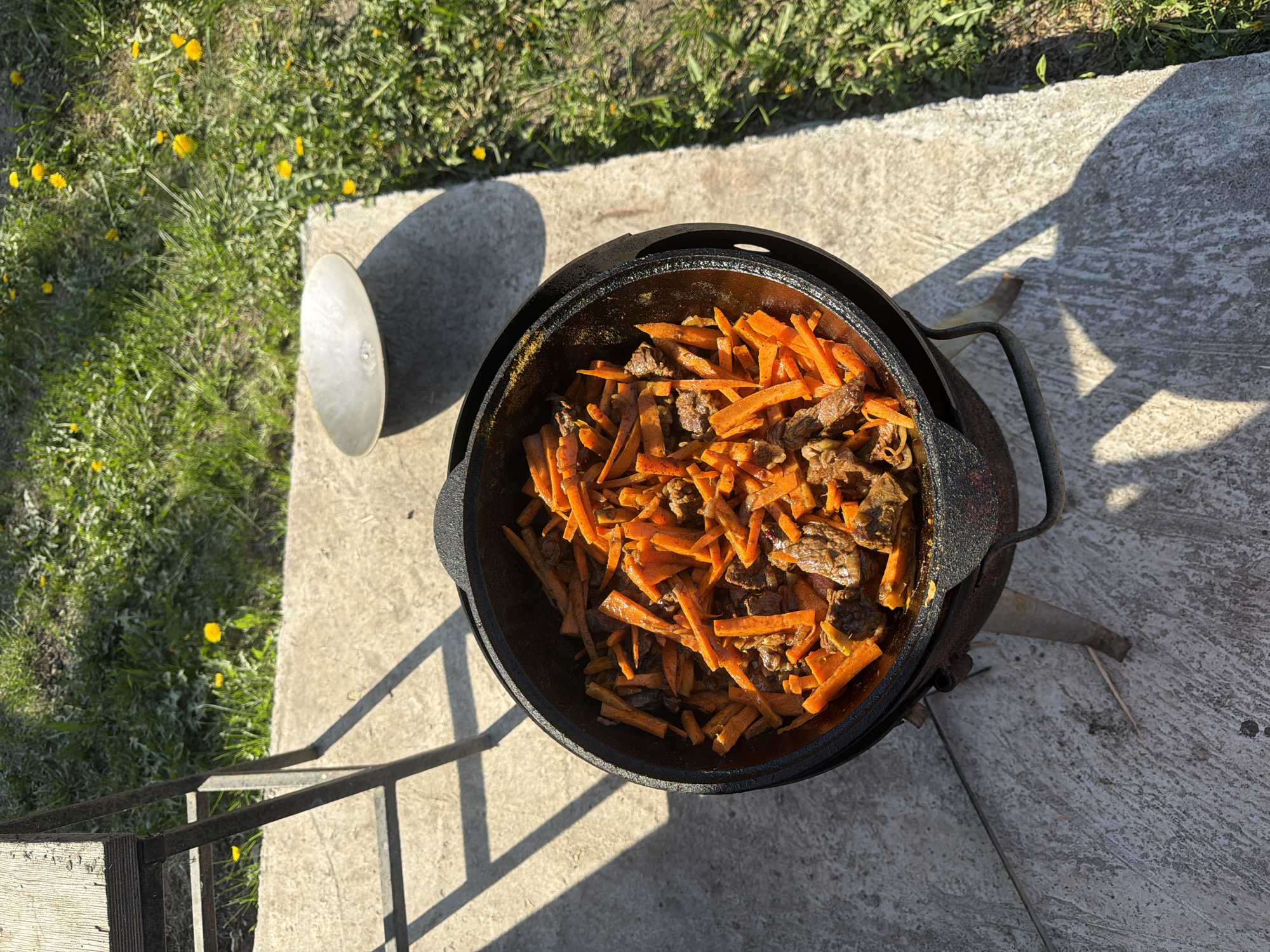 The image shows a large, black, cast-iron cauldron resting on a concrete surface. Inside the cauldron is a dish consisting of what appears to be carrots and meat. A lid rests on the ground beside the cauldron. The surrounding environment includes grass with yellow flowers and a metal railing, possibly indicating a patio or outdoor cooking area. The location is identified as Большая Шильна, Russia.