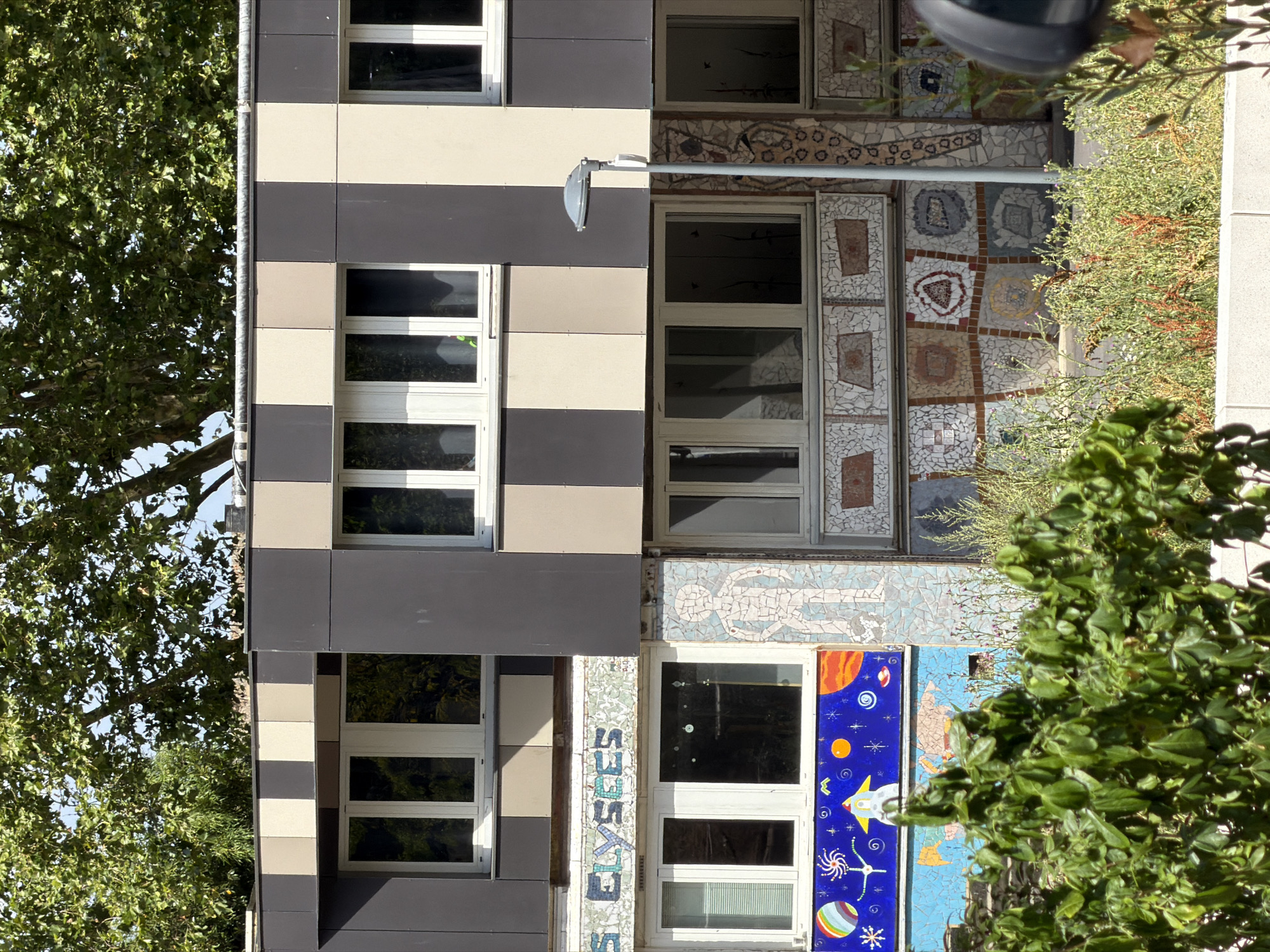 This is a vertical, medium shot of a modern building facade in Évry-Courcouronnes, France. The building features a striking design with alternating bands of dark gray and cream-colored panels. Interspersed among these panels are numerous windows with white frames. The facade is further embellished with colorful mosaic tile artwork.
To the left of the building, a large, leafy green tree with dense foliage partially obscures the view of the windows. To the right, the building's mosaic art becomes more prominent, featuring abstract patterns and depictions of celestial bodies, including planets, stars, and a rocket ship against a vibrant blue background. Some of the mosaic work appears to be in a grid pattern with square inserts. There is also a close-up view of vertical mosaic text that appears to read "ÉLYSÉES."
A single outdoor light fixture is mounted on a horizontal pole attached to the building. The scene is illuminated by natural daylight, suggesting it is daytime. The overall impression is one of an artistically designed, contemporary urban building with elements of nature and space-themed art.