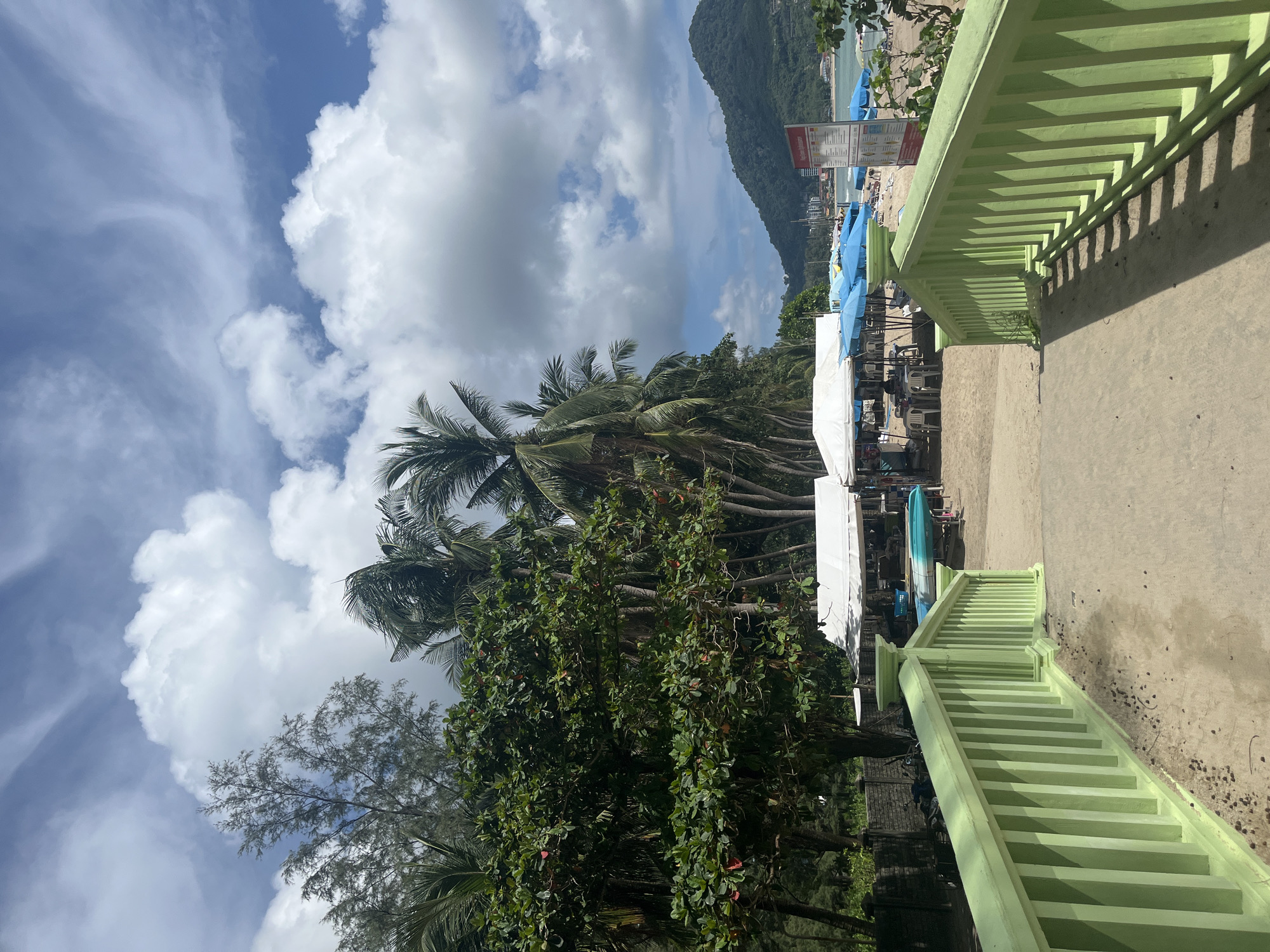This is a photograph taken at a beach in Ban Bang Thao, Thailand. In the foreground, a light green railing is visible, leading down a concrete path. Beyond the railing, the sandy beach stretches out, dotted with white tents and numerous blue umbrellas, suggesting a market or food stalls. Palm trees and other lush green vegetation line the edge of the beach, and in the background, a verdant hill rises. The sky is a vibrant blue, with dramatic white clouds. A few people are visible under the tents, seated at tables. The overall impression is of a sunny day at a tropical beach resort.