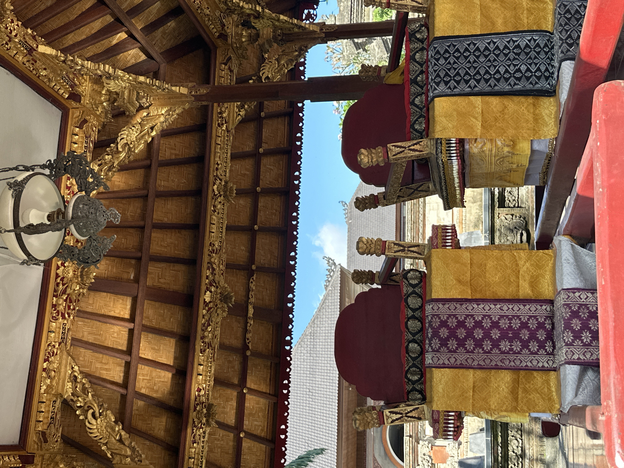 The image captures a detail of a traditional Balinese temple structure in Ubud, Indonesia. The focal point is two ornate, unoccupied thrones, upholstered in red with decorative gold accents and fabric coverings in yellow and purple, or yellow and black. These thrones are situated beneath a meticulously crafted wooden roof structure adorned with intricate gold carvings and a woven bamboo ceiling. A vintage chandelier hangs from the ceiling. In the background, traditional Balinese temple architecture with grey tiled roofs and carved stone elements is visible under a bright blue sky. The overall impression is one of cultural richness and ornate craftsmanship.