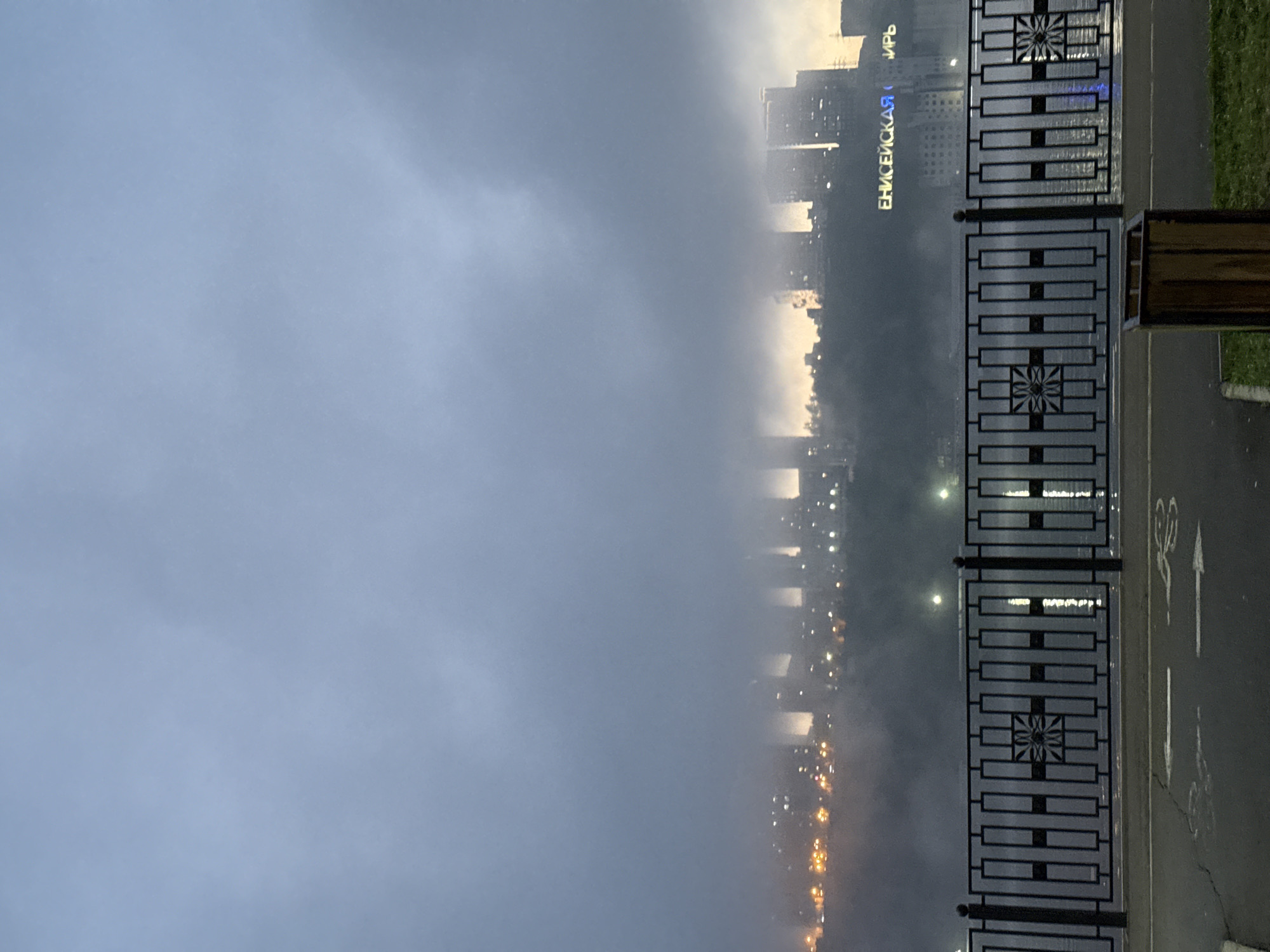 The image shows a foggy urban scene in Krasnoyarsk, Russia. A black metal fence with geometric designs runs along the right side of the frame, next to a paved path with a bicycle lane marking. Beyond the fence, a river is visible, obscured by heavy fog. On the opposite side of the river, the city skyline is faintly visible through the fog, with building lights and illuminated signage in Cyrillic letters, "ЕНИСЕЙСКАЯ" (Yeniseyskaya), indicating the location. The sky above is overcast, with the top right suggesting a break in the clouds, perhaps from the sun.