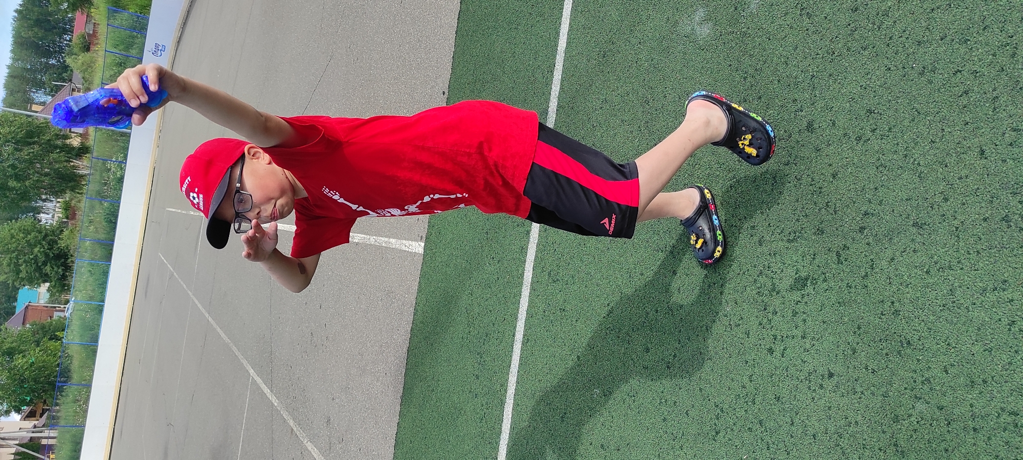 A young boy in a red t-shirt, black and red shorts, a red baseball cap, and glasses is holding a blue water gun aloft with his right arm outstretched. He is standing on a green artificial turf surface with white lines marking a court. He is looking upwards and to his left. His left hand is raised to his forehead, as if shielding his eyes or in a salute. In the background, there is a grey paved court, a white barrier, a blue fence, and trees and houses. The image was taken in Noviy, Russia.