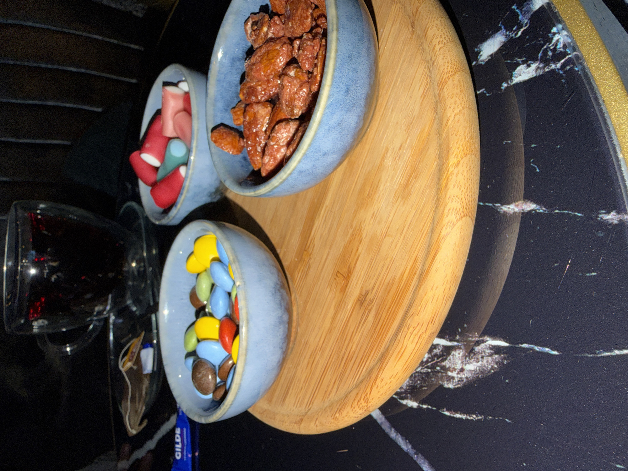 A wooden tray holds three blue ceramic bowls filled with snacks. The bowl on the left contains colorful candy-coated chocolates. The bowl in the middle holds pieces of red and light blue gummy candy. The bowl on the right is filled with candied nuts, likely almonds or pecans. Behind the bowls, a glass mug filled with a dark liquid, possibly tea or coffee, sits on a saucer. A blue wrapper with the word "GILDE" is visible on the left side of the image. The tray and bowls are placed on a dark, marbled surface. The scene appears to be indoors, with dim lighting. The exact location in Nagold, Germany, cannot be determined from the provided image.