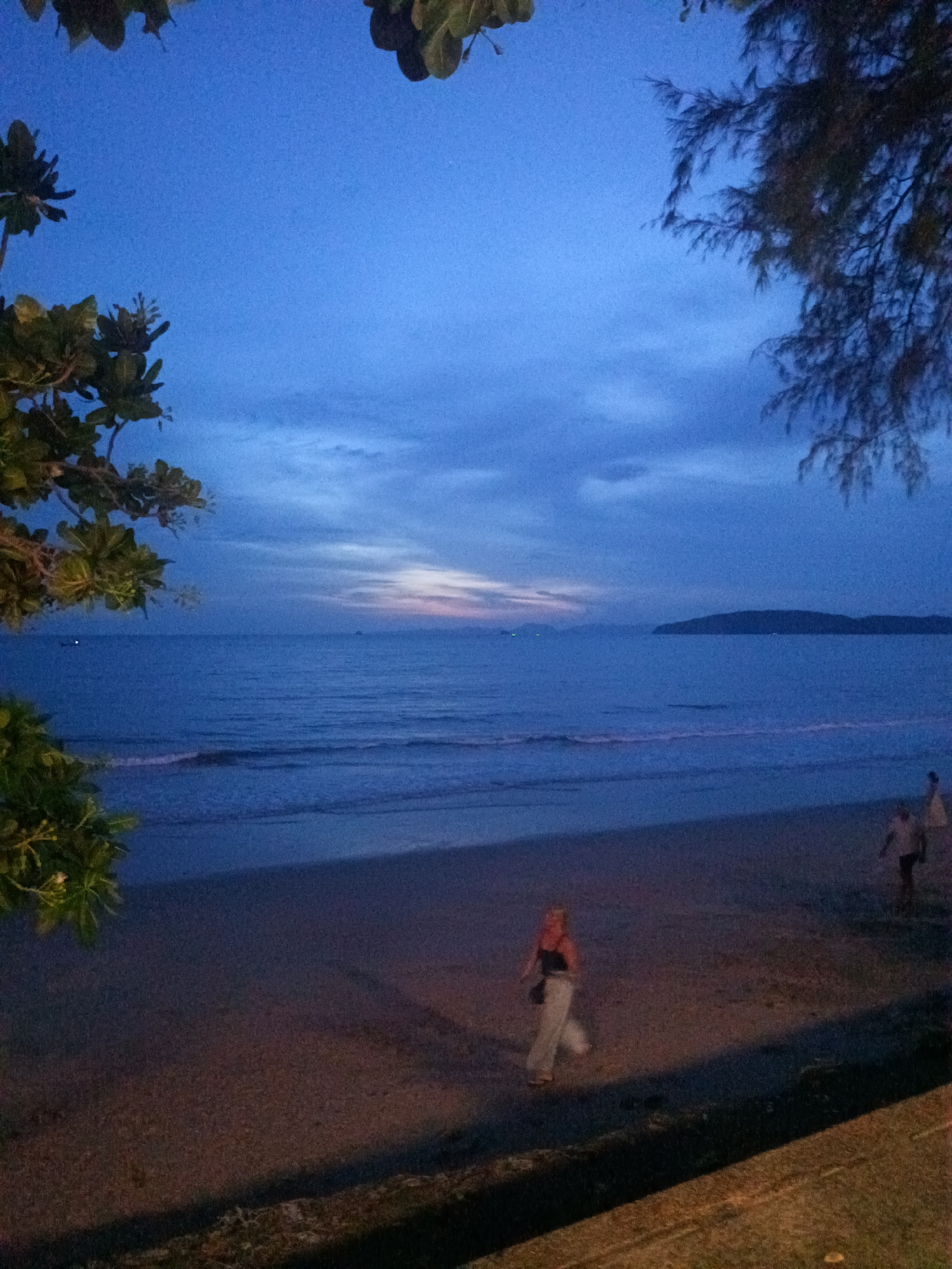 The image shows a beach at dusk in Ban Ao Nang, Thailand. The sky is a deep blue with some orange hues visible on the horizon. The sea is calm with gentle waves reaching the shore. In the foreground, there is a sandy beach with two individuals walking. The beach seems to be slightly illuminated by artificial light. There are trees on the left and right of the frame, partially obscuring the view of the sky. An island is visible in the distance across the water.