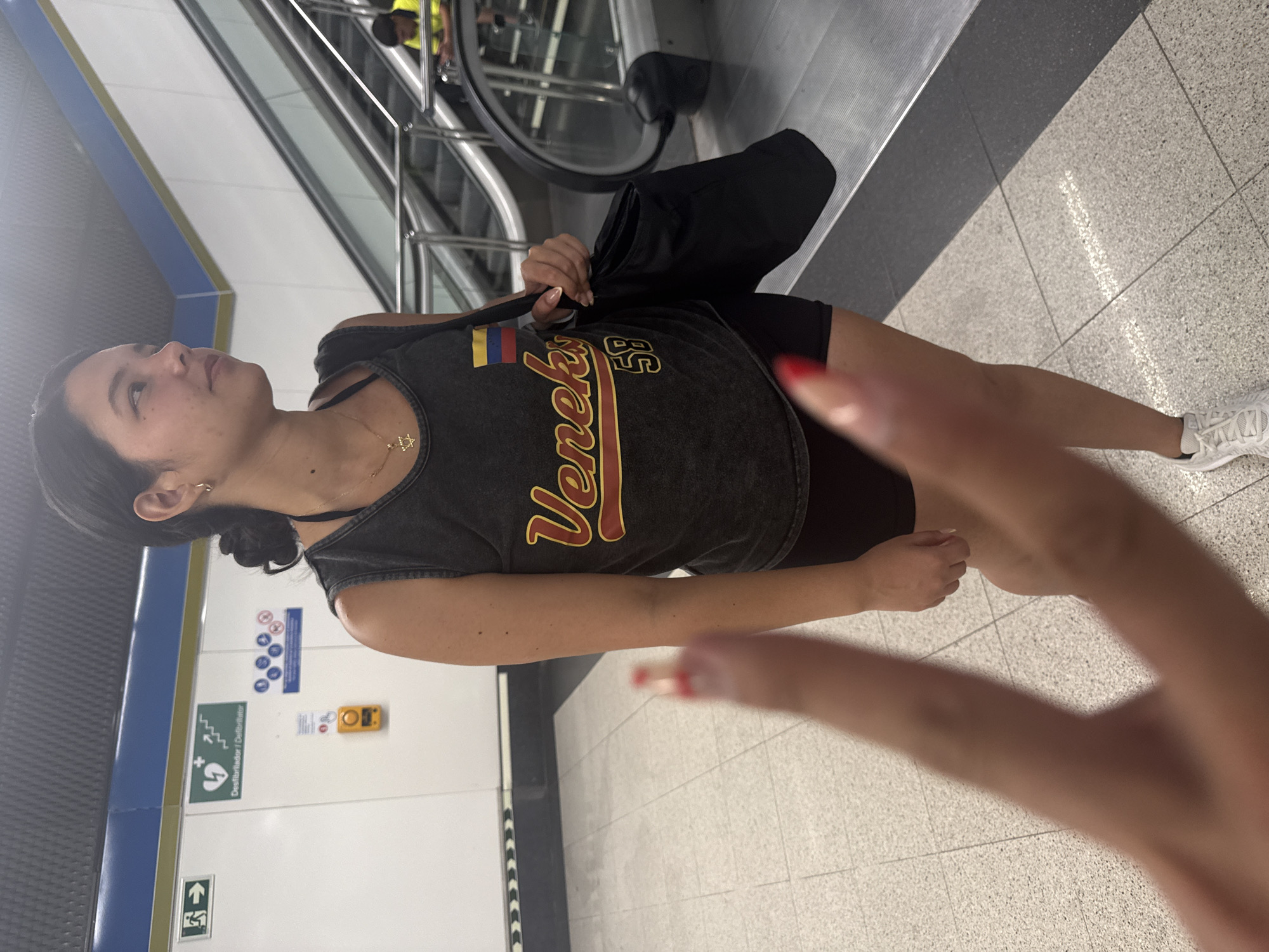 The image shows a young woman standing in a subway station. She is wearing a dark tank top with "Veneko 58" printed on it and black shorts. She has a black tote bag slung over her shoulder and is looking to her right. A blurry finger in the foreground is positioned to look like it is pointing at her. In the background, an escalator is visible, with another person in a yellow vest at the bottom. The walls of the station are white with blue and yellow trim, and there are signs indicating emergency exits and a defibrillator. The floor is tiled with a light gray and white speckled pattern. The image appears to have been taken in Getafe, Spain.