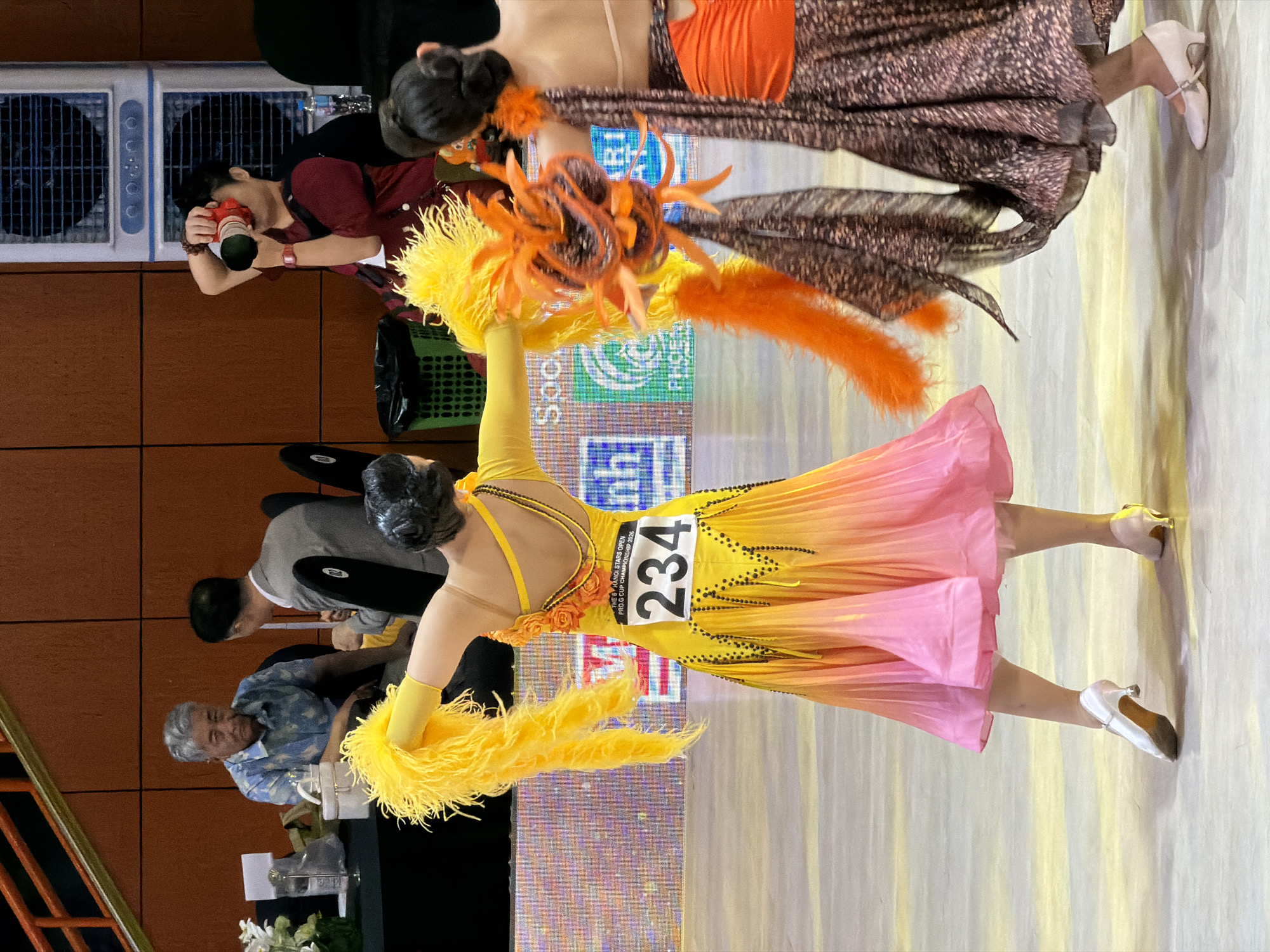 This image depicts a ballroom dancing competition, likely held indoors, with a focus on two female dancers in elaborate costumes. One dancer, in the foreground and facing away from the camera, wears a vibrant yellow and pink ombre dress adorned with feathers and beadwork. A bib with the number "234" is visible on her back. She has yellow feather accents on her arms. To her right, another dancer, also facing away, wears a brown patterned dress with an orange accent piece on her torso and orange feather details.
In the background, several spectators are seated. An older man with grey hair, wearing a blue patterned shirt, is on the left. Next to him, another man in a grey suit is also seated. Further back, a person in a dark red top is holding a camera and appears to be taking a photo. Two large white air conditioning units are mounted on a wooden wall in the background. A green bin is also visible. The floor appears to be a polished surface reflecting light. The context suggests a formal event, possibly an international competition given the presence of a photographer and spectators. The specific location within Hà Nội, Vietnam cannot be determined from the visual information alone.