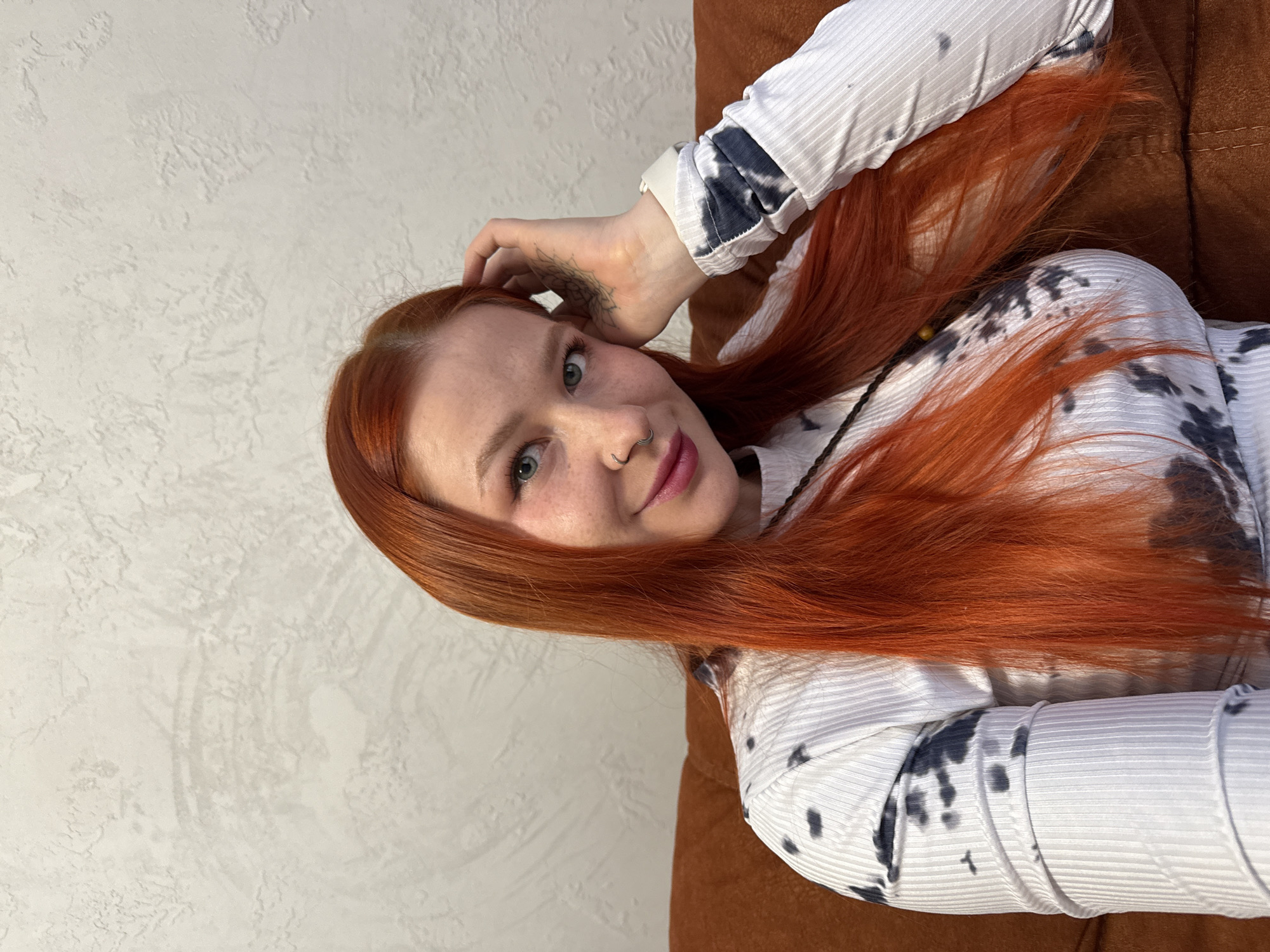 A female subject is depicted from the chest up, seated indoors in Rostov-on-Don, Russia. She has long, straight, vibrant red hair and light-colored eyes. Her complexion is fair with visible freckles across her nose and cheeks. She has a silver hoop piercing in her left nostril and her lips are painted a shade of pink. The subject wears a long-sleeved, ribbed top that is predominantly white with irregular dark grey tie-dye patterns. Her right hand is raised, with fingers touching the side of her head and hair; a tattoo featuring a floral or mandala design is visible on the palm side of her right wrist/hand. She maintains direct eye contact with the viewer and exhibits a slight smile. A thin, dark cord with small beads is partially visible around her neck. The background consists of a light grey, textured wall and an orange-brown upholstered surface, likely a sofa or armchair, upon which the subject is seated.