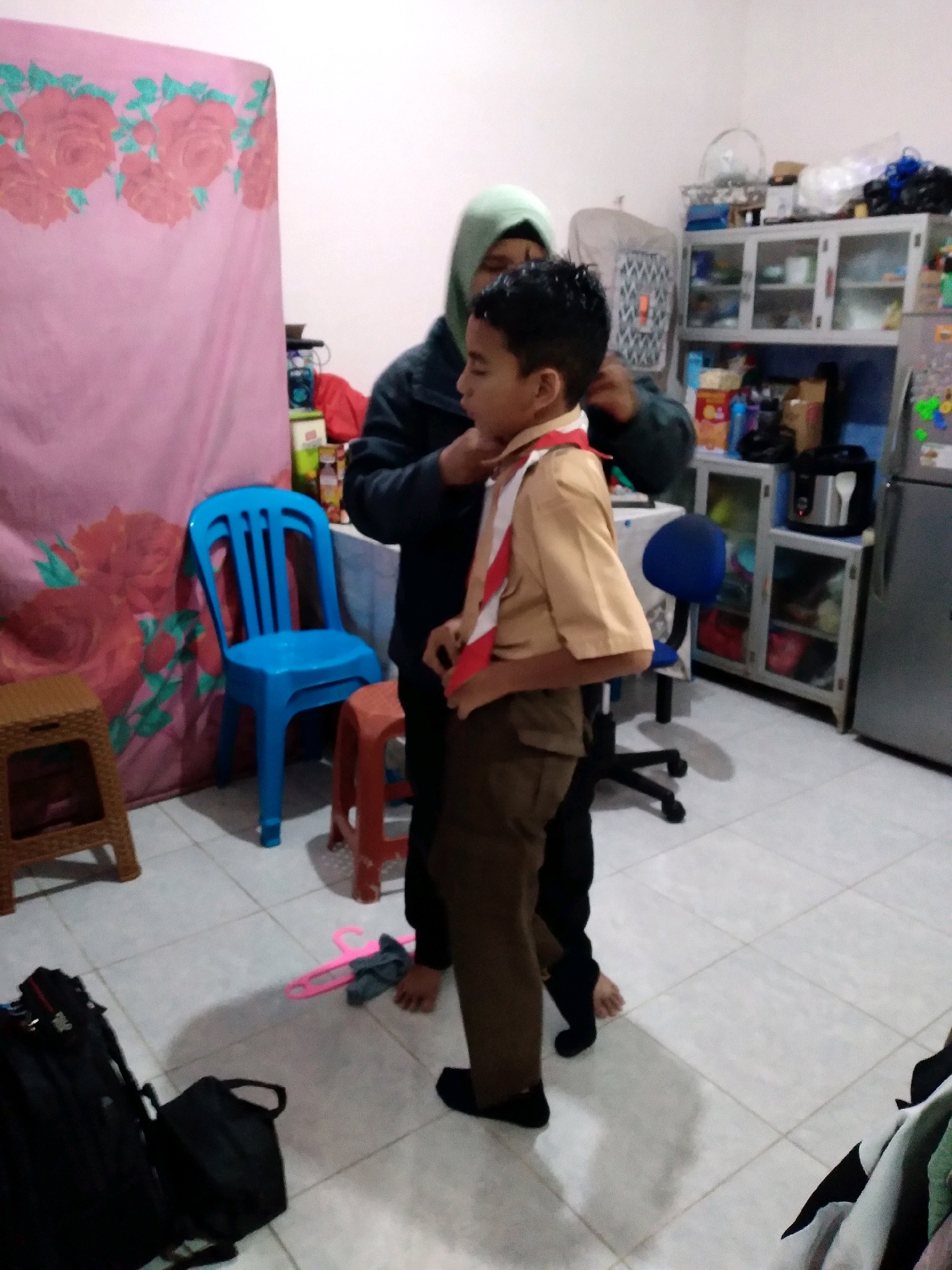 In Cimahi, Indonesia, a young boy in a scout uniform, consisting of a tan shirt, brown pants, and a red and white sash, is being helped with his attire by an adult, possibly a parent or guardian, who is partially obscured. The scene takes place indoors, within a room with tiled flooring. To the left, a pink fabric backdrop with a floral pattern is visible, along with a small wooden stool. A bright blue plastic chair stands prominently in the center-left. In the background, a shelf unit filled with various items, including food containers and appliances, is present, alongside a silver refrigerator. A black backpack and another bag are on the floor to the left. A pink plastic hanger and a dark-colored garment are also on the floor in the foreground. The lighting suggests an indoor environment with artificial light sources.