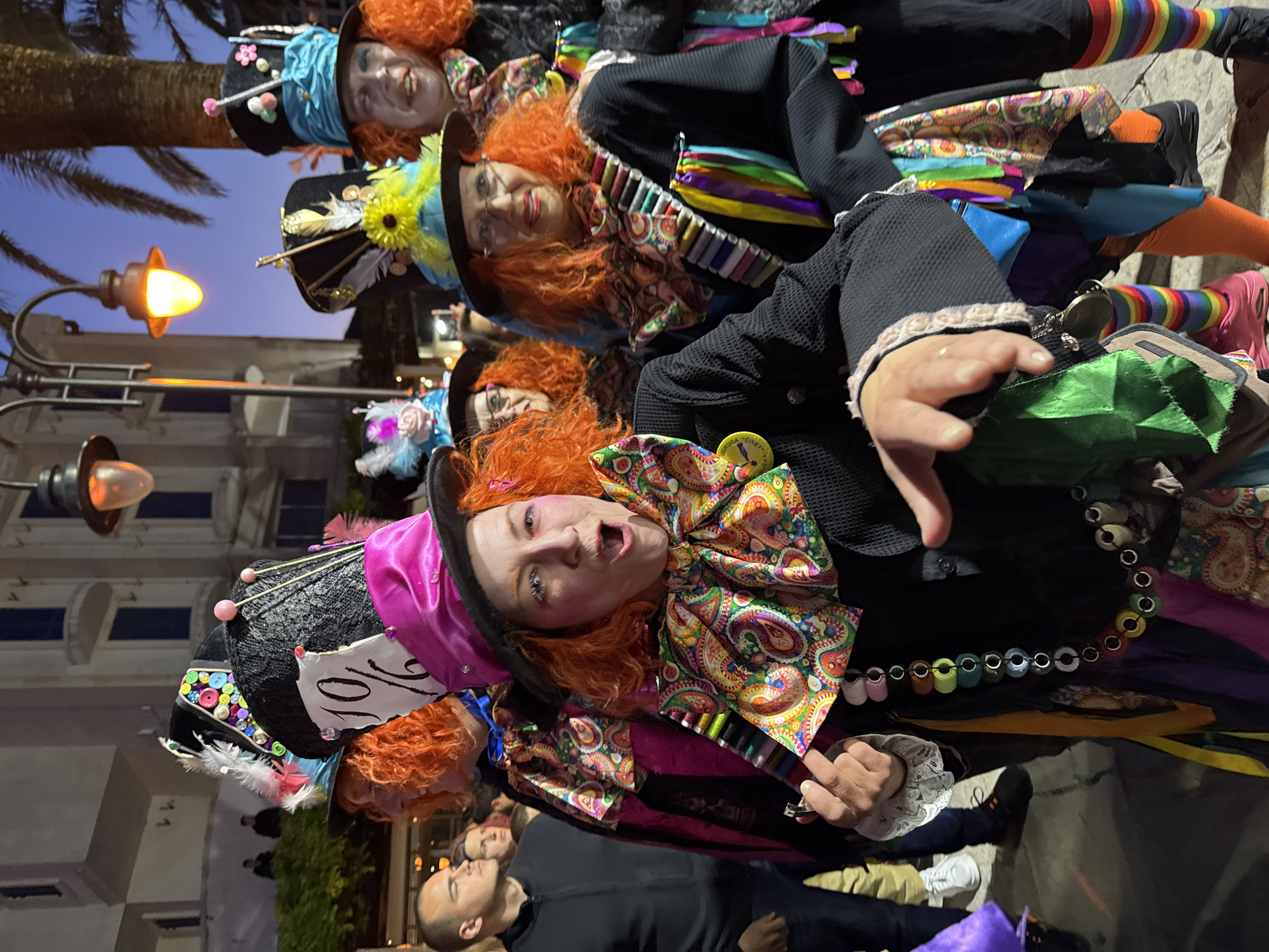 The image captures a vibrant evening scene in Budva, Montenegro, featuring a group of individuals in elaborate, colorful costumes, primarily resembling the Mad Hatter from Alice in Wonderland. The scene is set outdoors, likely on a street or promenade, indicated by visible palm trees and streetlights, with a dark blue twilight sky overhead.
The main subjects are several people, mostly women, dressed in black jackets with highly decorative and mismatched elements. They wear bright orange, curly wigs and distinctive top hats adorned with various items such as pins, ribbons, feathers, buttons, and fabric swatches. One hat clearly displays the "10/6" tag, a classic Mad Hatter reference. Their attire also includes large, colorful paisley bow ties, striped sleeves or leggings, and layered fabrics in a kaleidoscope of hues. One person's costume features a belt-like adornment made of colorful spools of thread or buttons.
The individuals are expressive; one person in the foreground has their mouth open as if speaking or singing and extends a hand towards the viewer, holding a green fabric bag. Their faces show makeup, contributing to the theatricality of their appearance. Behind them, other costumed figures are visible, some looking directly at the camera with smiles. In the lower part of the image, other people not in costume, including a man in a black shirt, are partially visible, suggesting a public event or gathering.
The activity appears to be participation in a themed event, parade, or carnival, given the intricate and coordinated costumes. The overall mood is festive and lively. Artificial light from street lamps illuminates the scene, creating warm glows against the fading natural light.