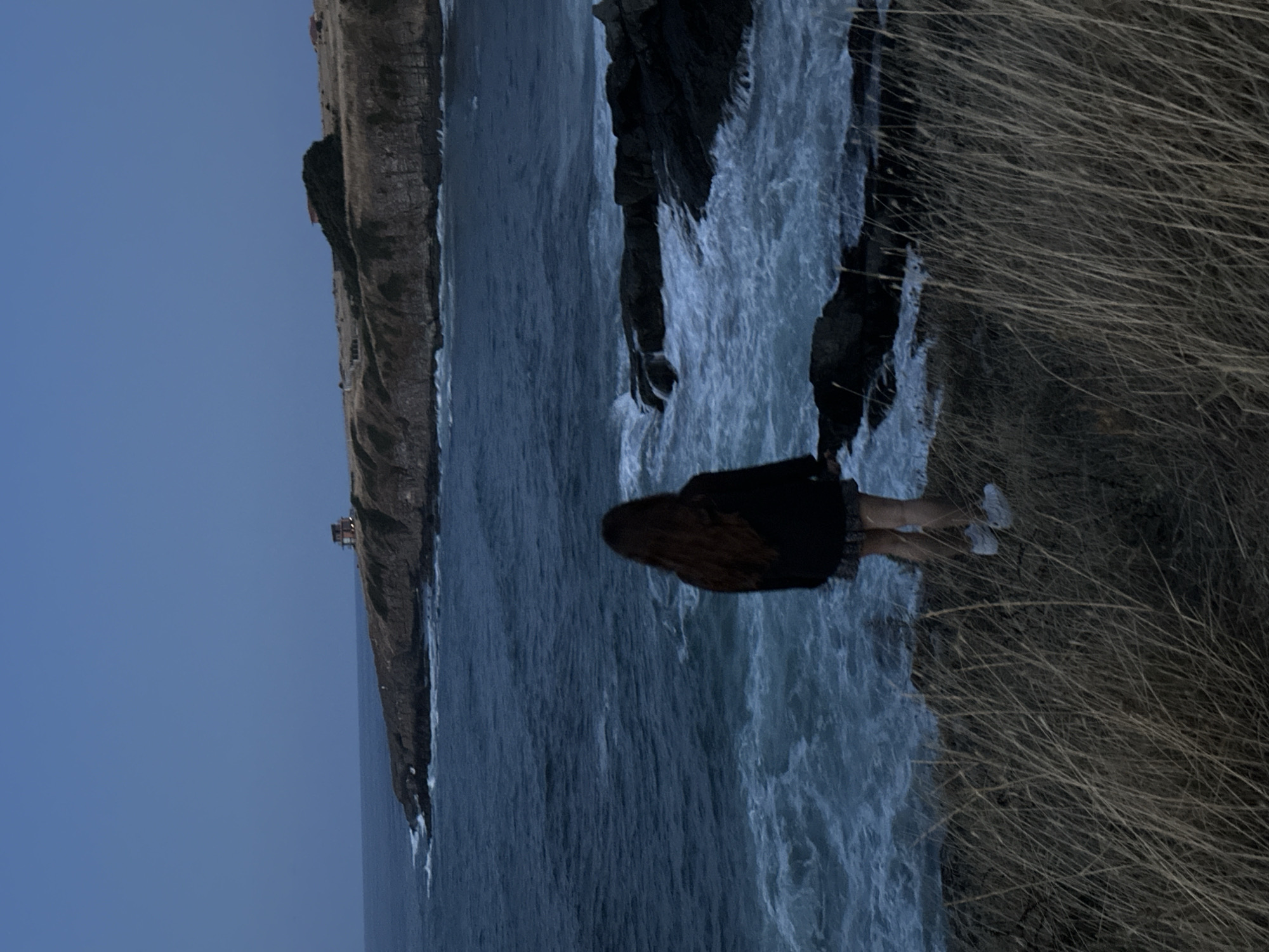 An overhead shot captures a young woman standing on a grassy cliff overlooking a choppy, dark blue sea. The woman, facing away from the camera, has long, dark hair and wears a black jacket and a short skirt. White sneakers are visible on her feet. The sea crashes against dark, jagged rocks below the cliff, creating white foam and spray. In the distance, a rugged coastline stretches out, with a few buildings perched on the bluffs under a twilight sky. The overall atmosphere is serene yet dramatic. This scene is likely located in Sinemorets, Bulgaria, known for its dramatic coastal landscapes and picturesque views.
