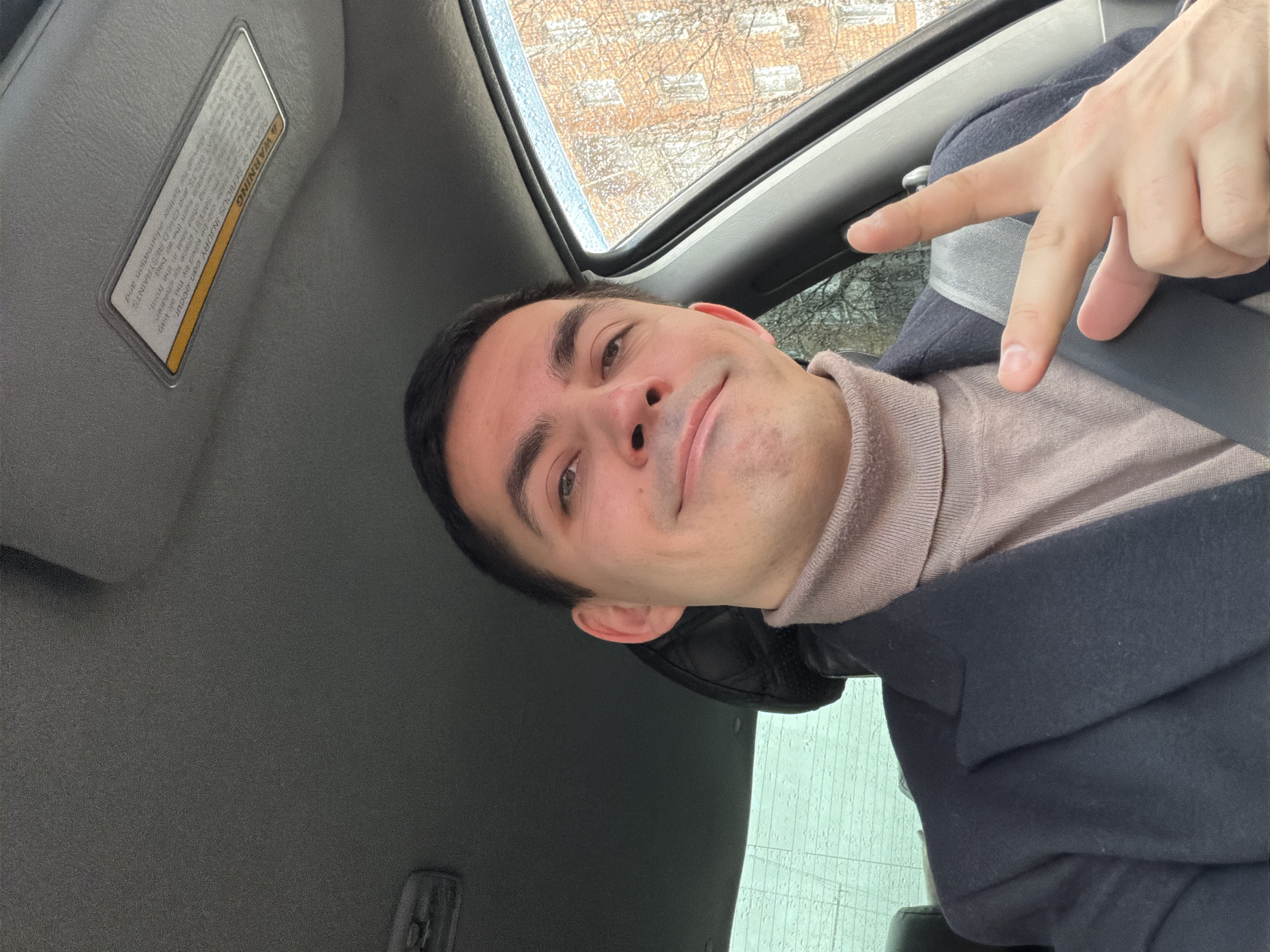 A man is featured in a selfie, taken from a low angle within the interior of a car. He appears to be a young adult with short dark hair and a clean-shaven face, looking directly at the camera with a subtle smile and a composed expression. He is dressed in a light beige turtleneck sweater layered under a dark blue or black coat or jacket. His right hand is visible in the upper part of the frame, making a peace or "V" sign with his index and middle fingers. He is wearing a seatbelt across his chest.
The car's interior is primarily gray, with a sun visor on the left side featuring a visible warning sticker. A sunroof above the man shows a blurred view of the outside, with what appears to be raindrops or moisture on the glass, suggesting wet weather. Through the sunroof, bare tree branches and brick-colored building facades can be seen, indicating an urban setting and potentially a colder time of year. The provided context states the location is Izhevsk, Russia.
The text on the warning label on the sun visor reads: "AWARNING DEATH OR SERIOUS INJURY can occur. and older can be killed by thi ac bag. Safe Manual for further information and SEAT BELTS and CHILD RESTRAINTS BACK SEAT SAREST place for children. and Facong child seat in the front. Date possible from the a bag."
