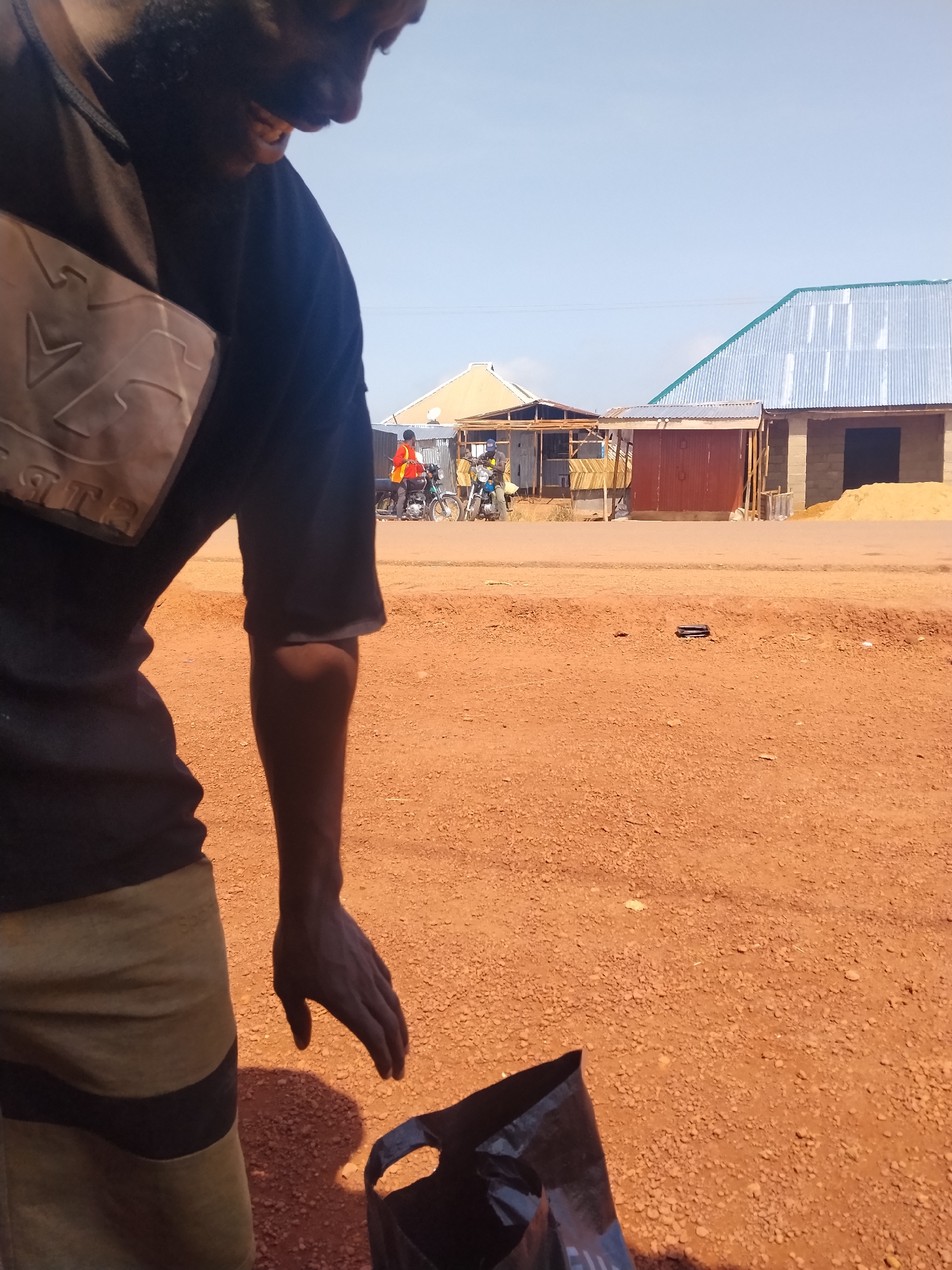 In Durumi, Nigeria, a person wearing a dark t-shirt and patterned shorts stands in an outdoor area with red, gravelly ground. The person's left arm is bent, and their hand is reaching down towards a black plastic bag on the ground. In the background, several buildings with corrugated metal roofs are visible, along with two motorcycles and two individuals. The sky is clear and blue.