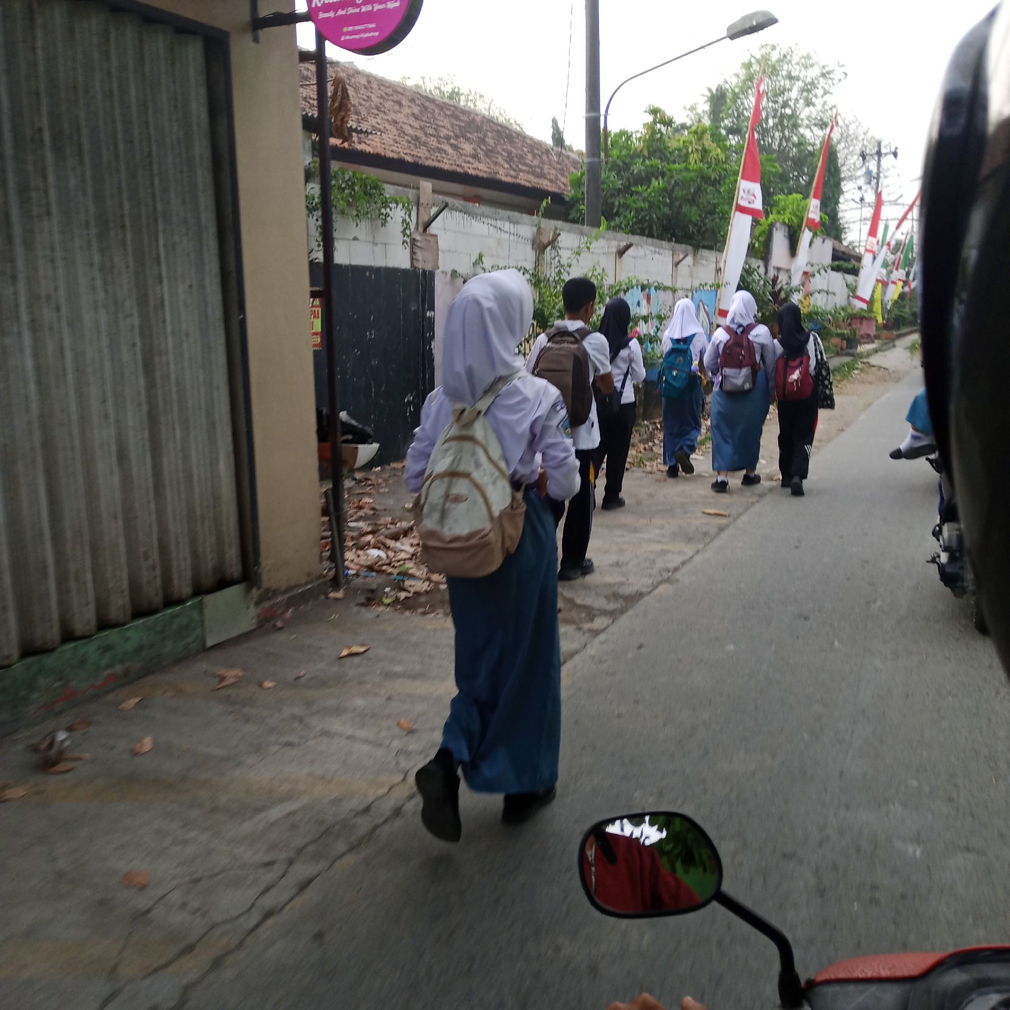 The image depicts a group of four students, two of whom are visible from behind, walking on a paved road in what appears to be a suburban or rural area in Katonsari, Indonesia. The students are wearing school uniforms consisting of white shirts and blue or black skirts, and most are wearing hijabs. They are carrying backpacks of various colors, including beige, blue, and red. One student is in the foreground, slightly ahead of the others, with a beige backpack. The students are walking away from the camera, heading down the road. The environment includes a sidewalk with some fallen leaves and debris, a building with a corrugated metal facade on the left, and a white wall with some graffiti in the background. In the distance, there are trees and flags on poles lining the road. The perspective suggests the image was taken from a moving vehicle, possibly a motorcycle, as indicated by the presence of a side mirror in the lower right corner of the frame, reflecting a red object and trees. The weather appears to be overcast, with a bright sky.