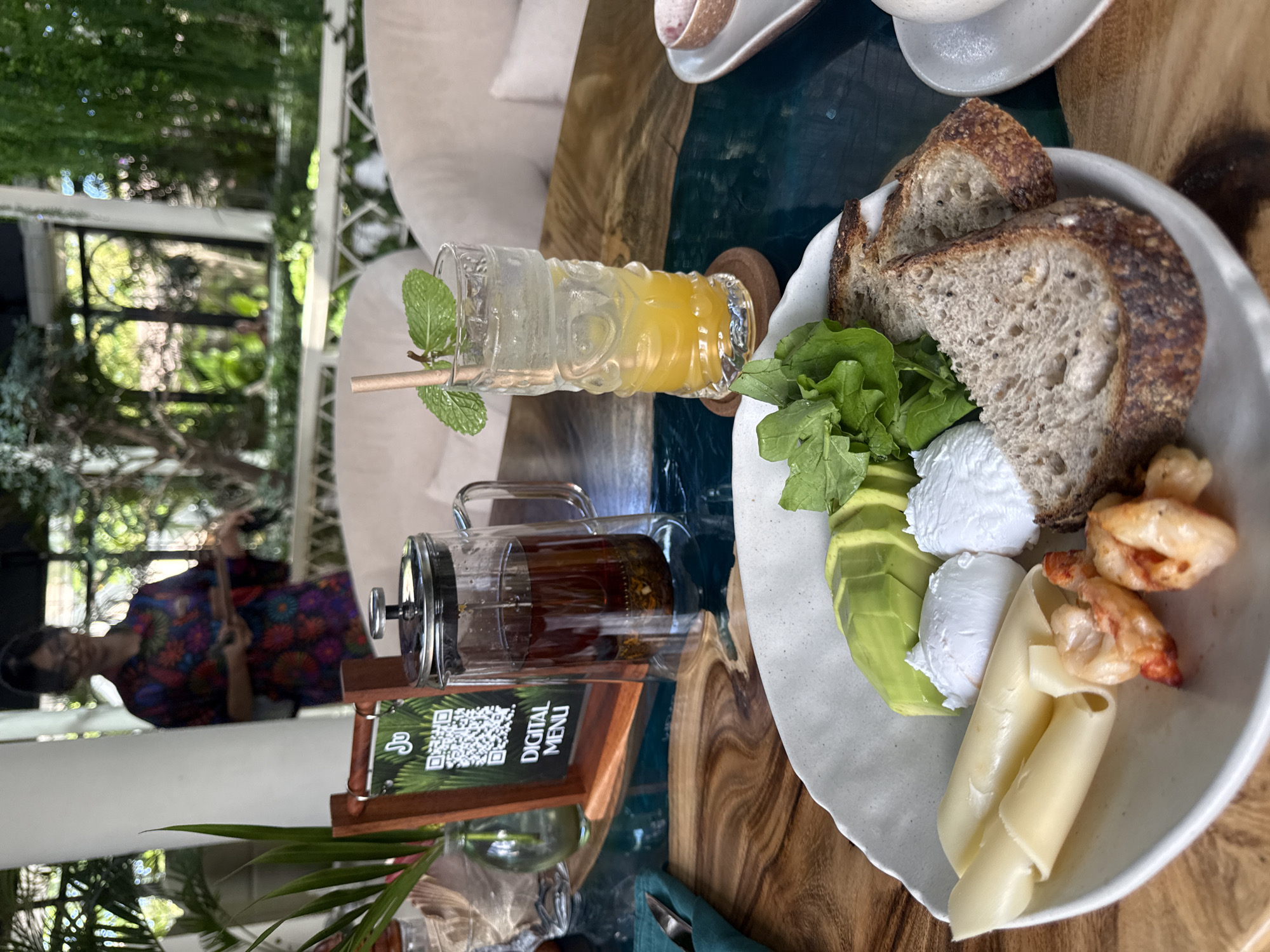 A table setting in North Kuta, Indonesia includes a white plate with sliced bread, greens, avocado, cheese rolls, and shrimp. A beverage with a mint garnish is nearby, along with a glass teapot and a QR code menu stand. A person wearing glasses and a patterned shirt stands in the background, and the location appears to be a cafe or restaurant with outdoor seating.