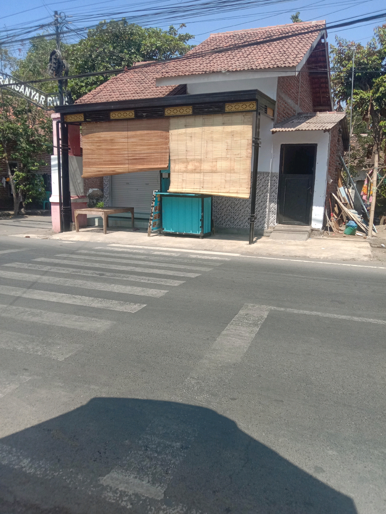 The image depicts a street scene in Karang Anyar, Indonesia, with a focus on a building that appears to be a shop or small business. In the foreground, a pedestrian crossing is visible on an asphalt road. The building to the right has a terracotta-tiled roof and a light-colored facade with a dark door. Two large bamboo blinds cover sections of the storefront. To the left of the building, a street sign that reads "RANGANYAR RW" is affixed to a utility pole. In front of the shop, a simple wooden table stands on the sidewalk. The environment is sunny, with a clear blue sky overhead, and trees are present in the background. There are no people or animals visible in the image. The scene appears to be quiet and somewhat desolate, suggesting it might be early in the day or that the business is closed.