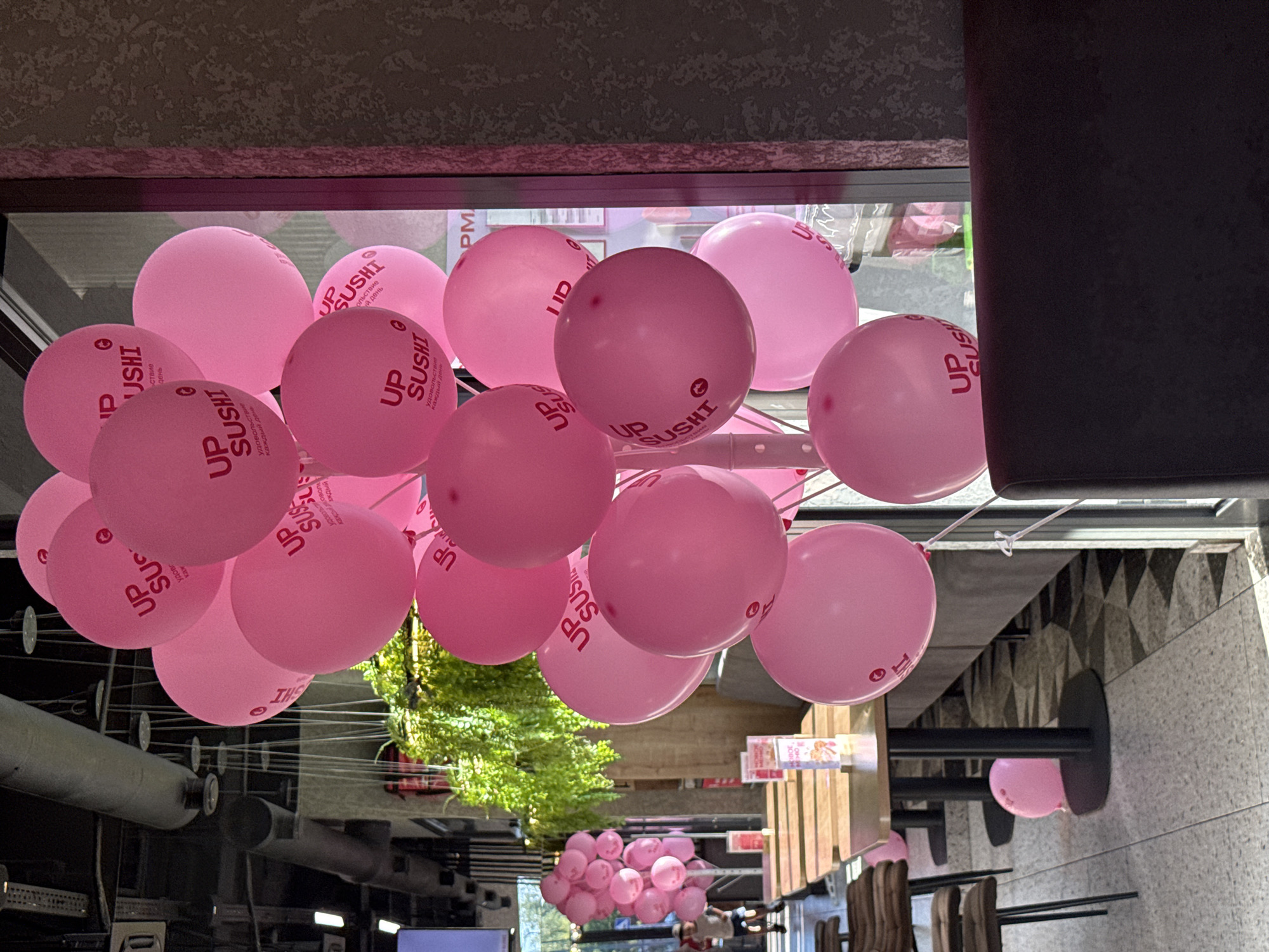 The image depicts a cluster of pink balloons, many of which are printed with the logo "UP SUSHI" and text in Russian. These balloons are prominently displayed indoors, appearing to be at a restaurant or event venue. In the background, tables and chairs are visible, suggesting a dining area. The lighting indicates it is daytime. One balloon is on the floor near a table leg. A faint reflection of a person is visible in the glass window on the right side of the image. Another cluster of pink balloons is visible in the background, to the left of the main cluster, with a person, likely a child, walking away from the camera. The overall setting is that of a commercial establishment decorated for a celebration or promotion.